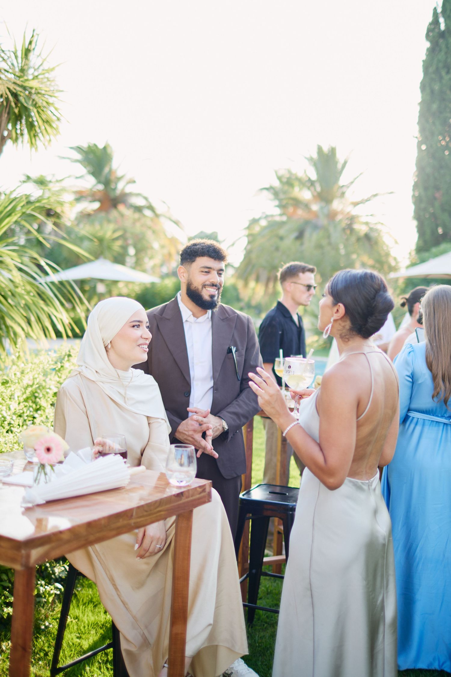 Wedding guests in elegant attire socialize at outdoor garden cocktail reception with palm trees in background.