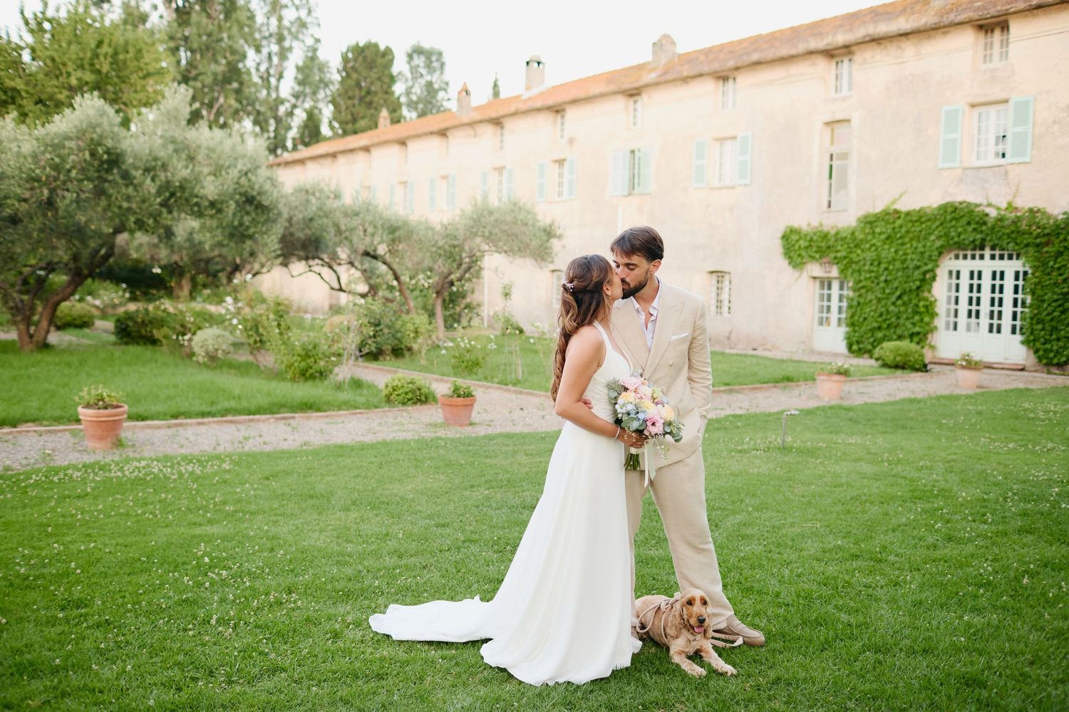 Bride and groom sharing romantic kiss with golden dog beside them on lawn at French chateau wedding.