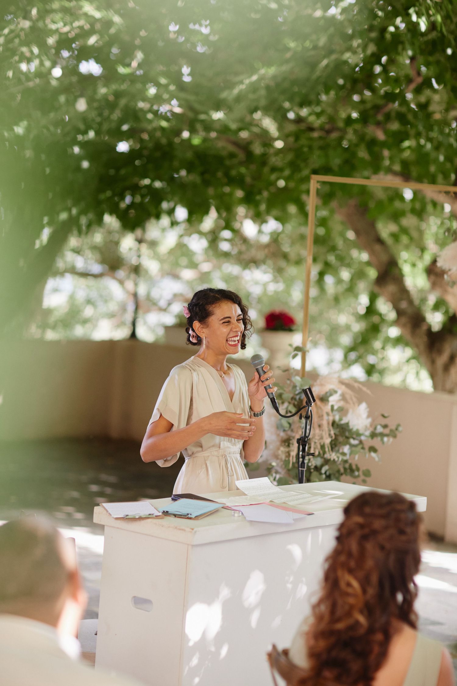 Joyful woman in white dress laughing while speaking at outdoor wedding ceremony podium under green trees.