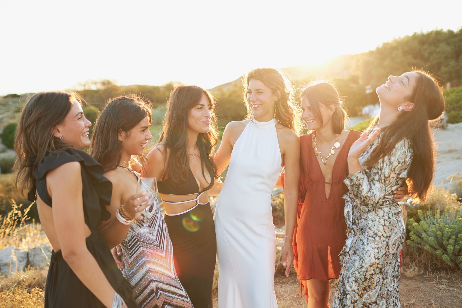 Bride in white halter dress laughing with bridesmaids at a golden hour outdoor wedding celebration.
