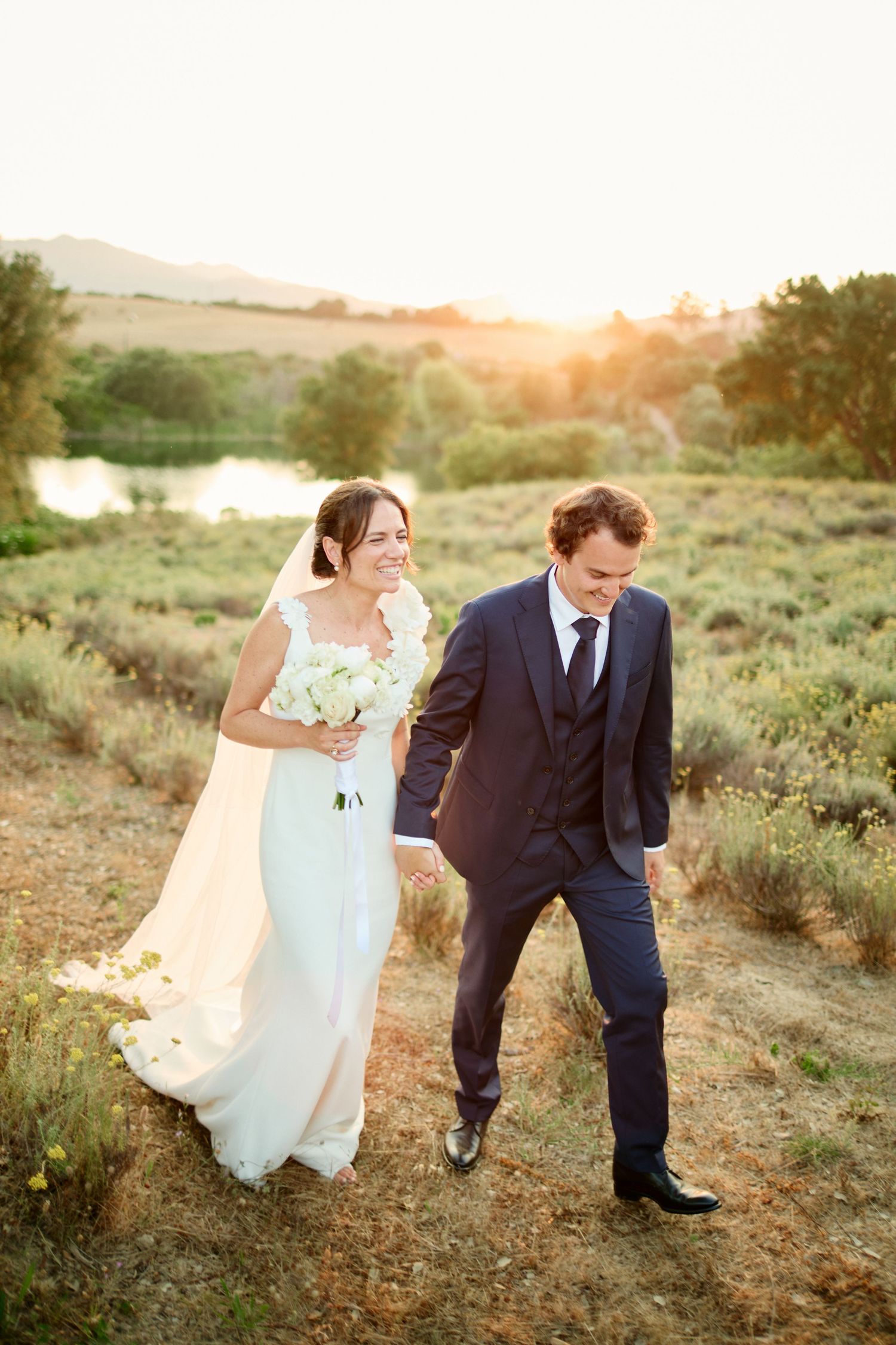Laughing bride in lace gown and groom in navy suit walk through golden sunlit countryside.