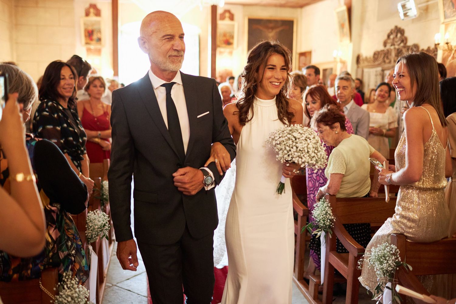 Bride in sleek white gown walking down church aisle with older gentleman escort, holding baby's breath bouquet.