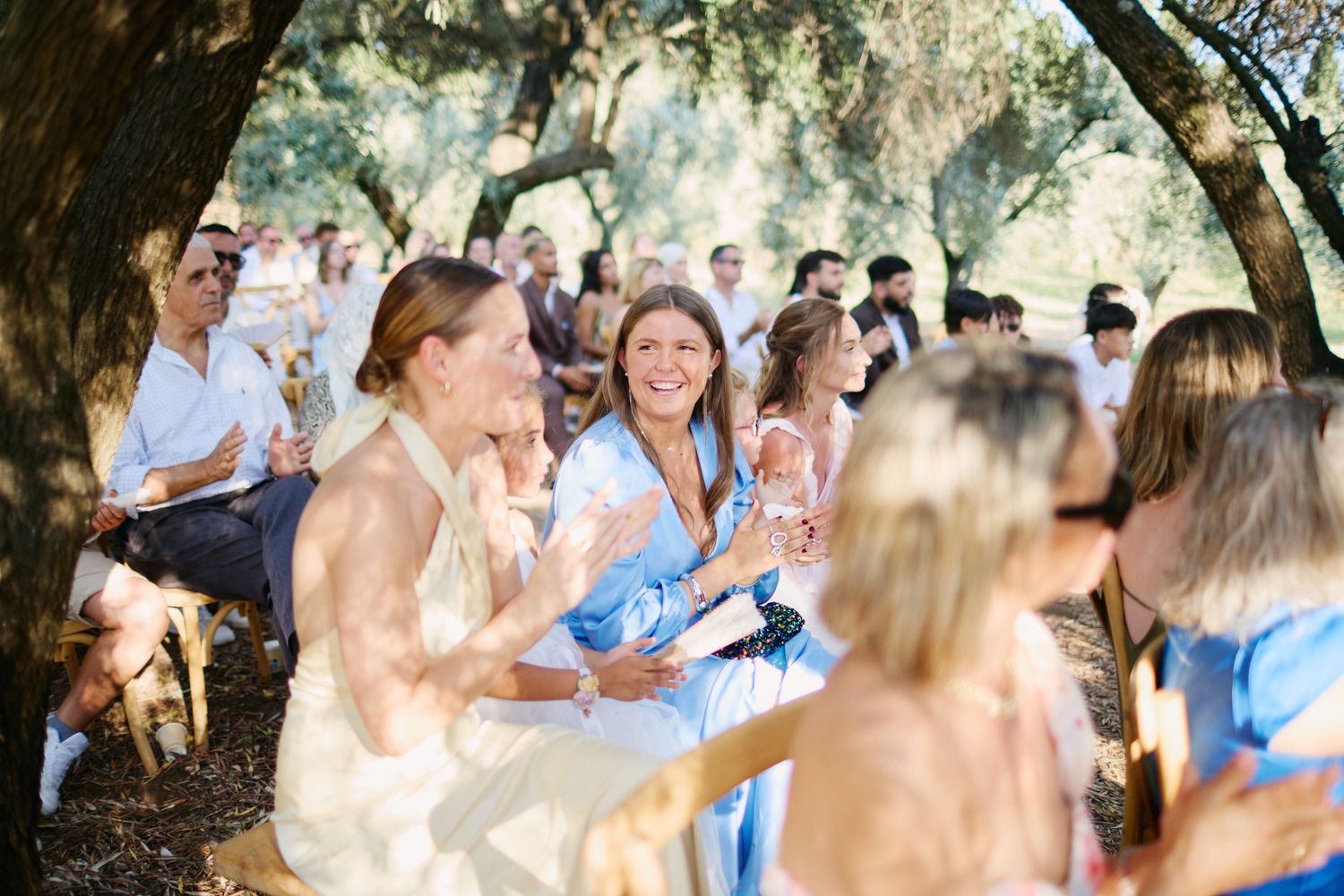 Smiling wedding guests seated outdoors under olive trees during a sunlit garden ceremony.