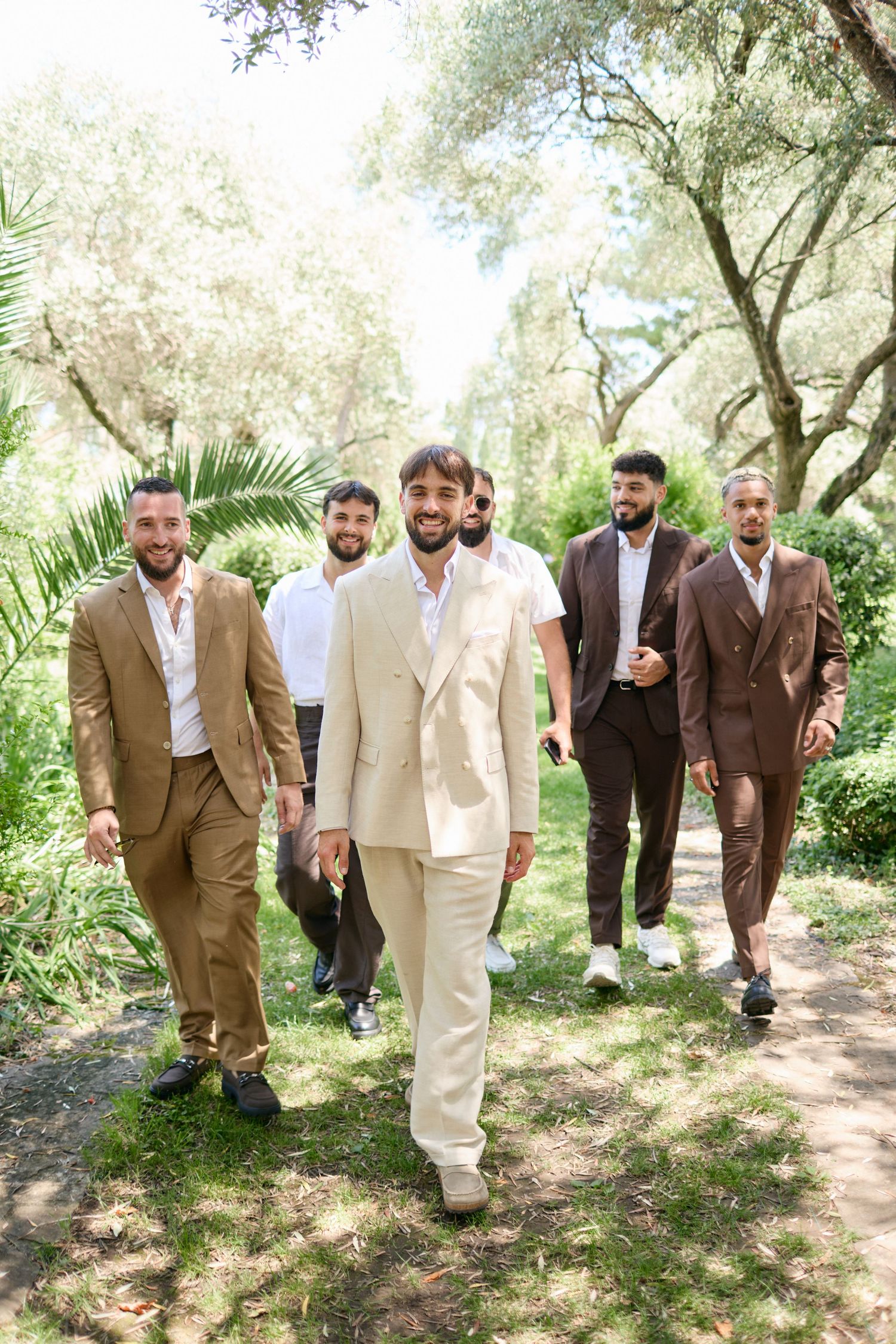 Smiling groom in cream suit leads groomsmen walking through a lush green outdoor garden path.
