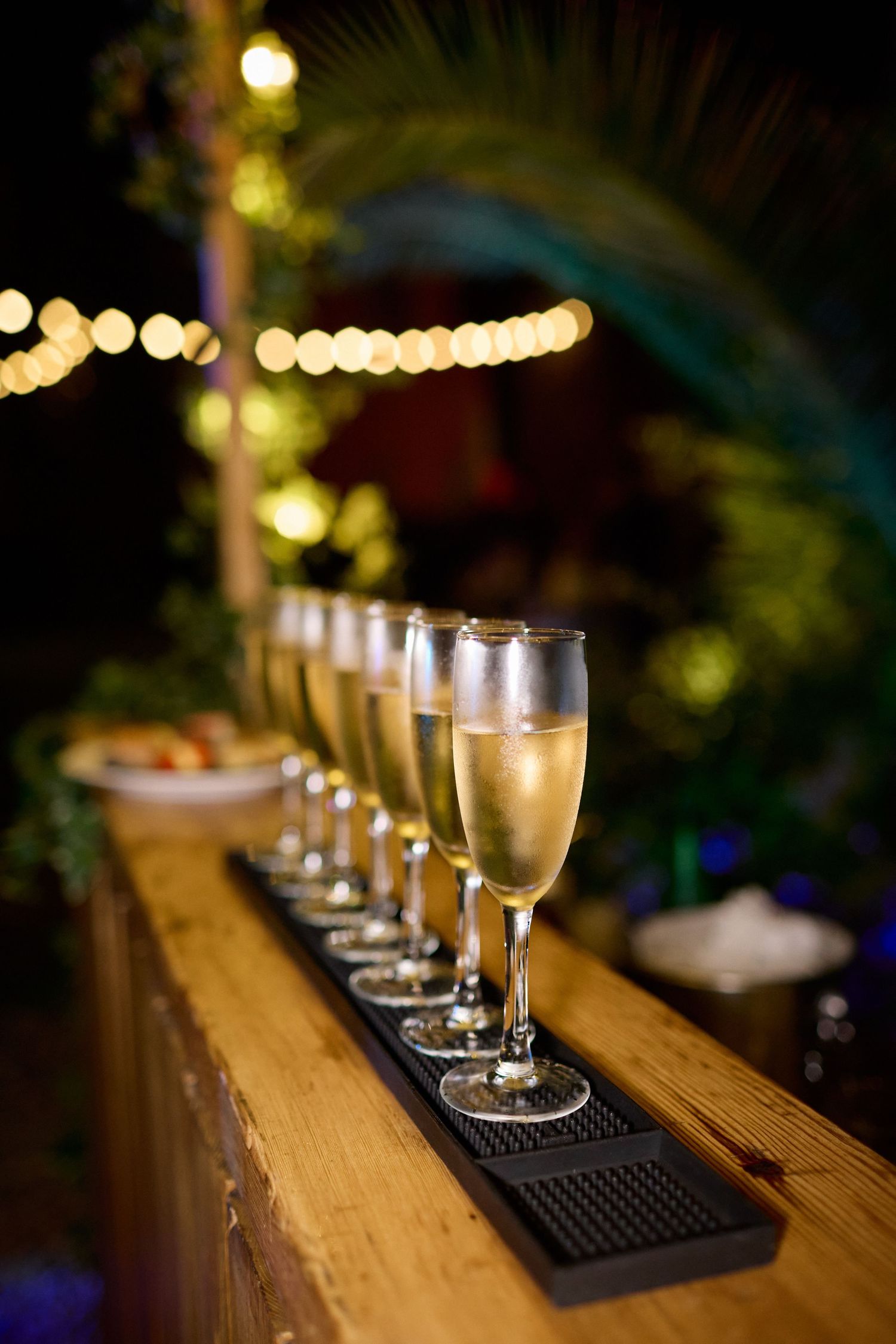 Row of champagne flutes lined up on a wooden bar at an outdoor evening wedding reception with string lights.
