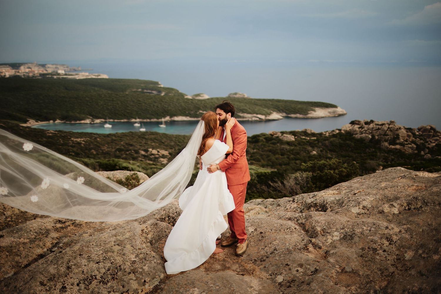 Wedding couple kissing on rocky coastal cliffs with bride's long veil dramatically blowing in wind.