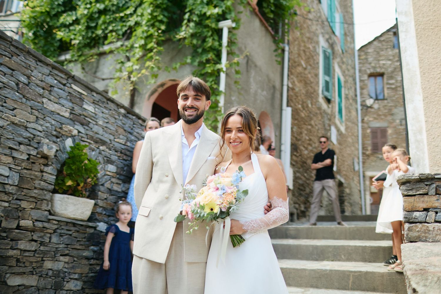 Newlywed couple poses smiling with bridal bouquet on rustic stone village steps after their ceremony.