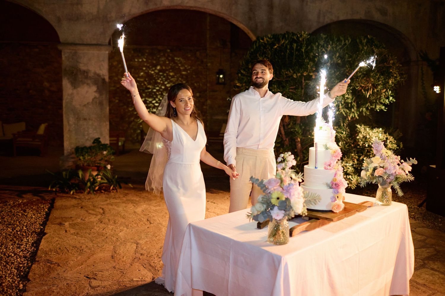 Bride and groom hold sparklers beside their floral wedding cake at a nighttime outdoor reception.