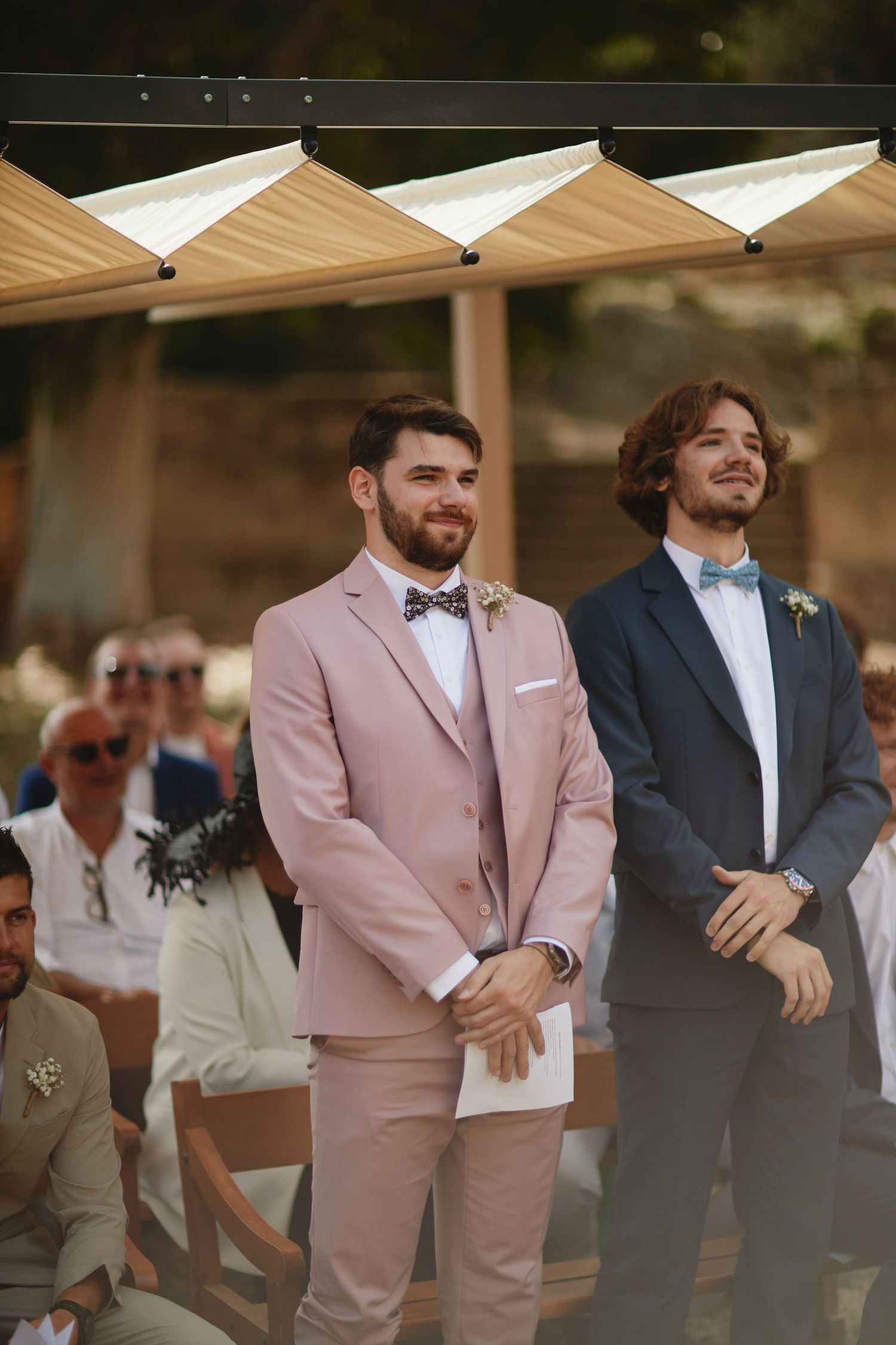 Two groomsmen standing at altar, one in pink suit with bow tie, other in navy suit at outdoor wedding.