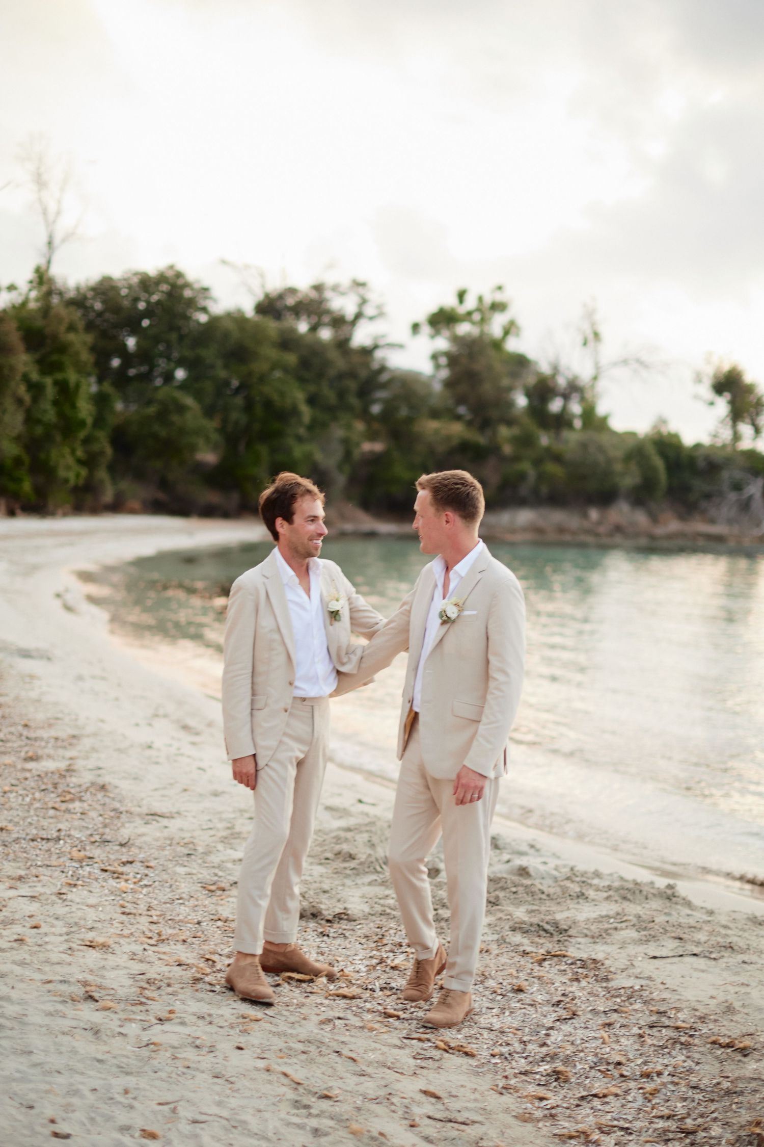 Two grooms in matching beige suits holding hands on a sandy beach at their wedding ceremony.