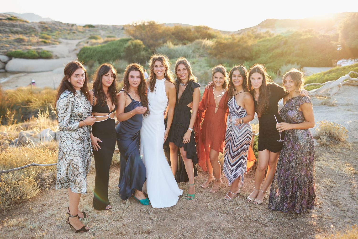 Bride in white gown posing with group of female wedding guests at sunset outdoor celebration.