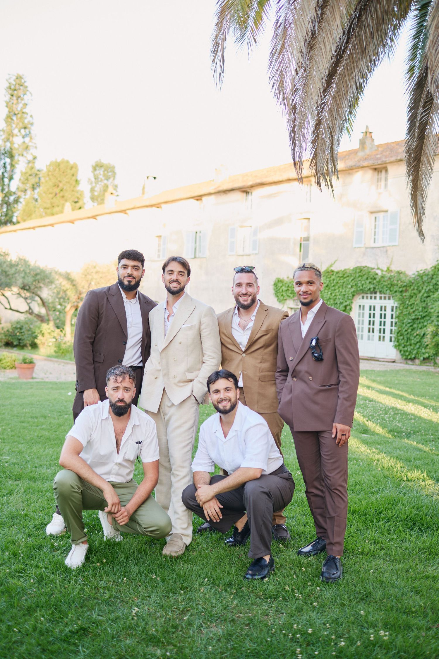 Six groomsmen posing in brown and beige suits on lawn by chateau.