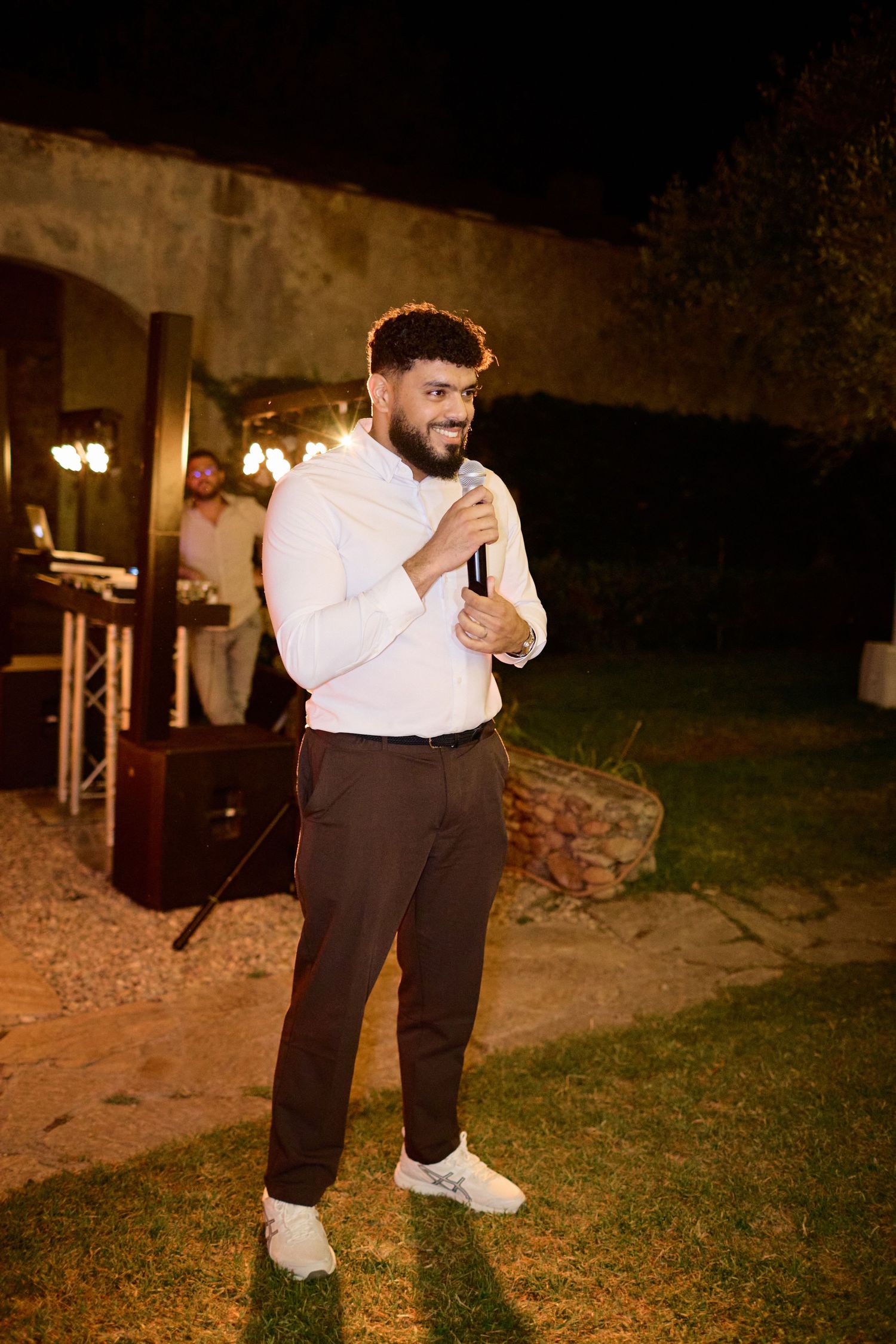Smiling man in white shirt delivers speech with microphone at outdoor evening wedding celebration.