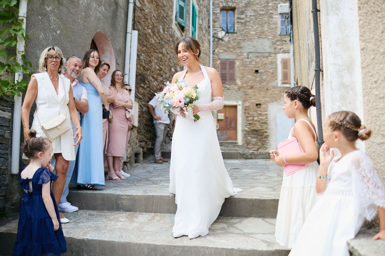 Elegant bride in white halter gown holds bouquet on stone steps surrounded by wedding guests and children.