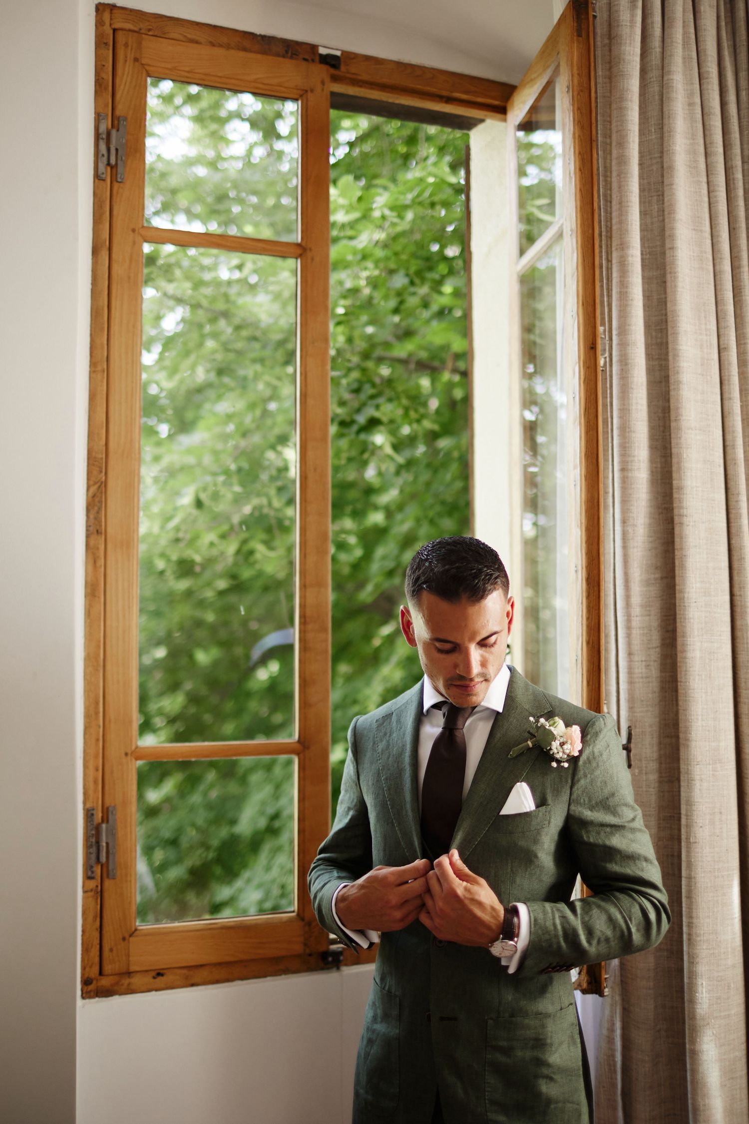 Groom in a green linen suit fastening his cuff links by an open wooden window with lush greenery outside.