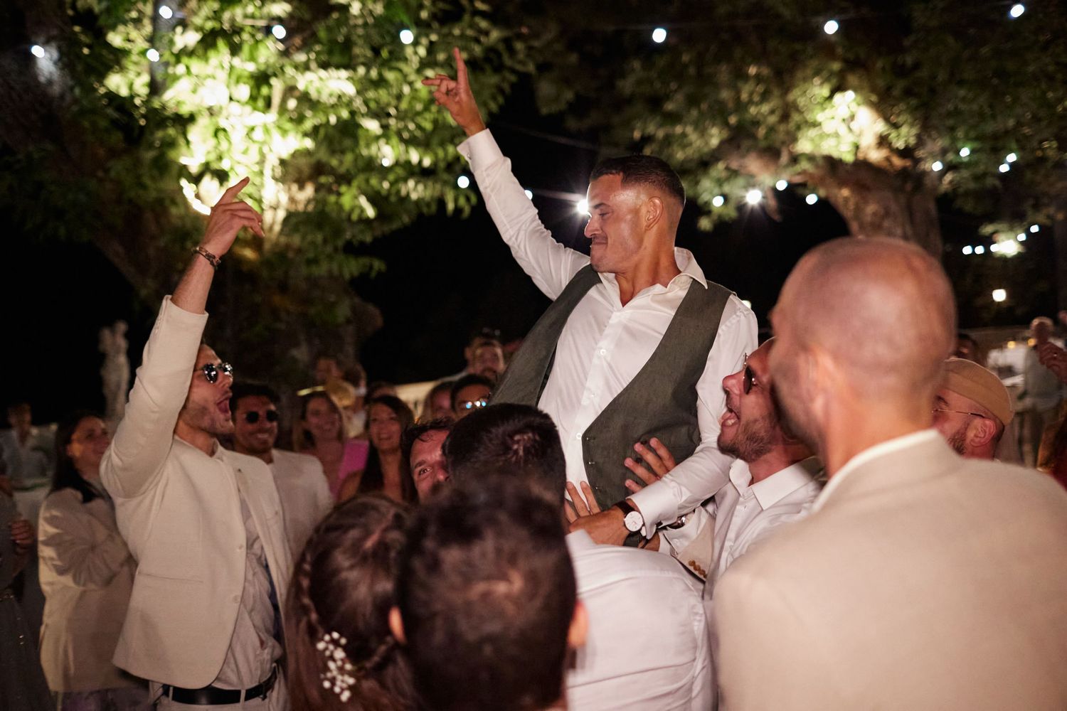 Wedding guest lifted by crowd during lively outdoor evening wedding reception celebration.