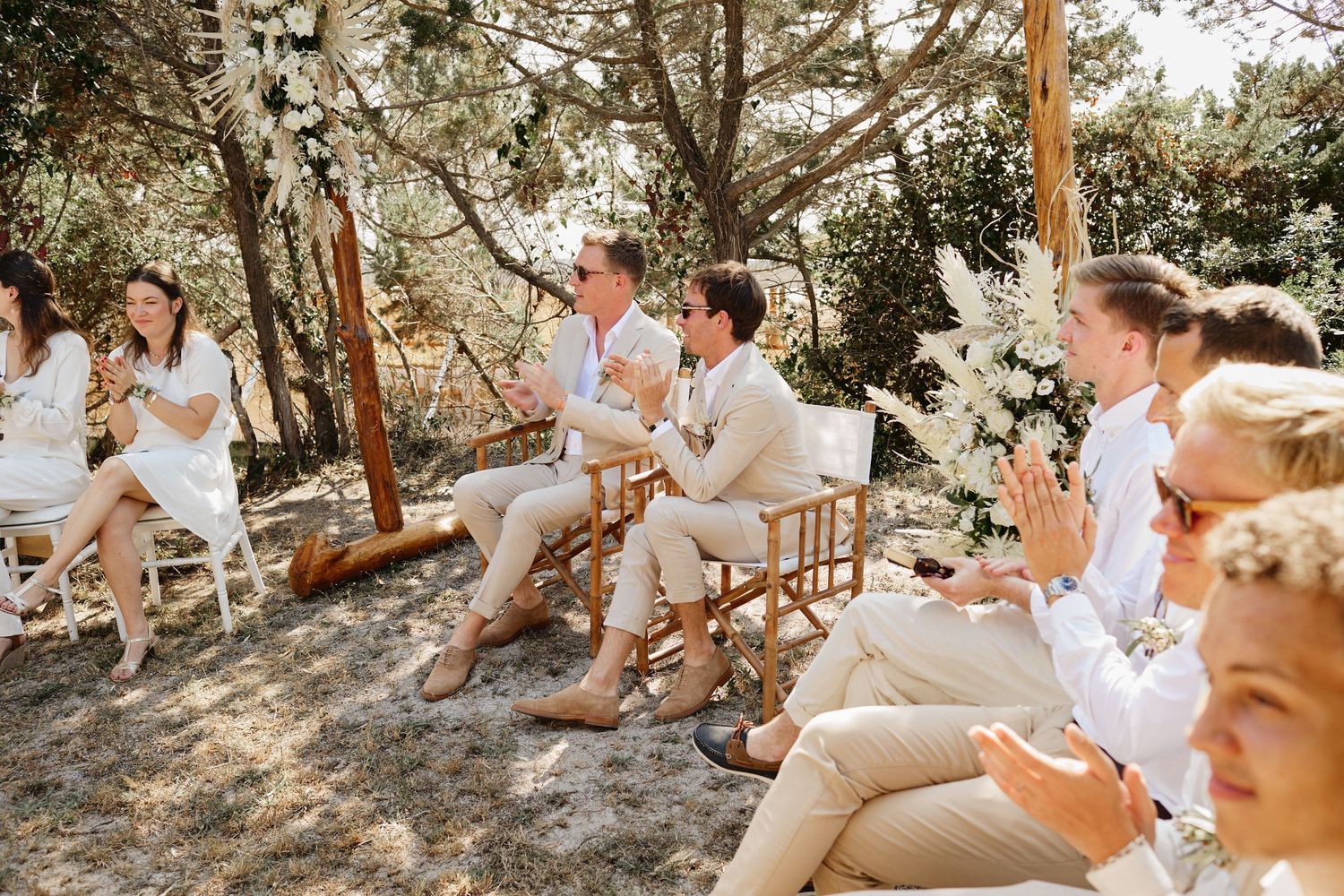 Guests in white and beige attire applaud during an outdoor wedding ceremony surrounded by trees.