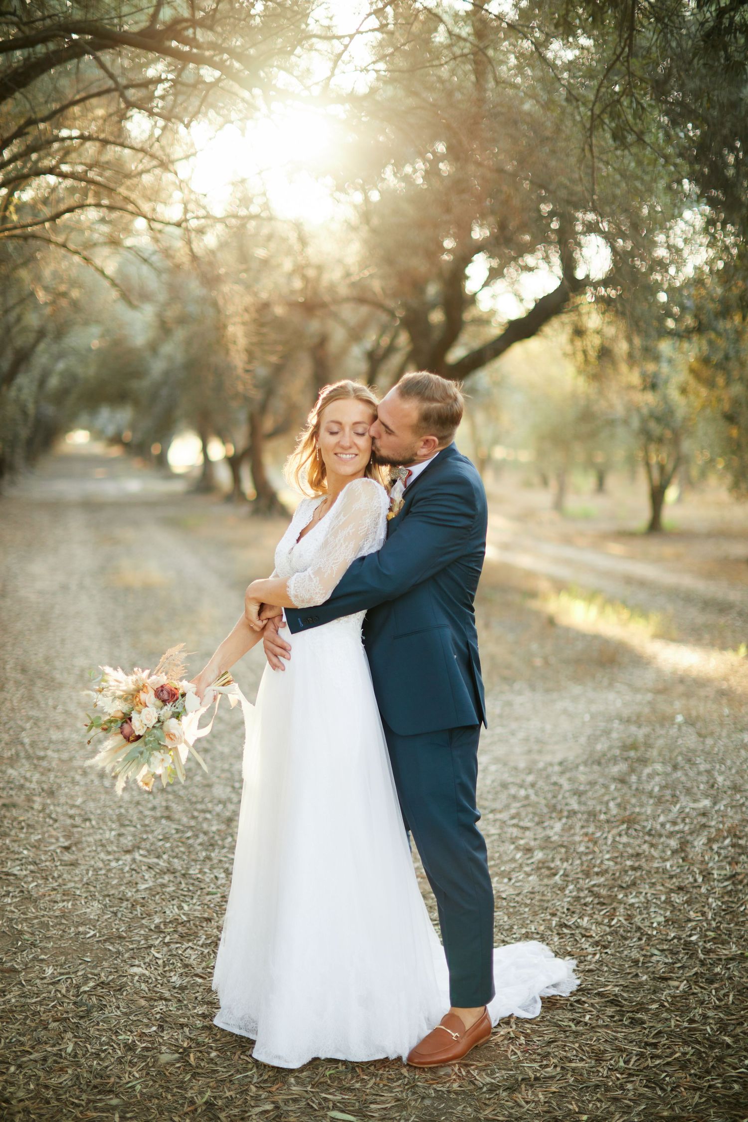 Wedding couple embracing in a sunlit olive grove, bride in white gown holding bouquet, groom in navy suit smiling.