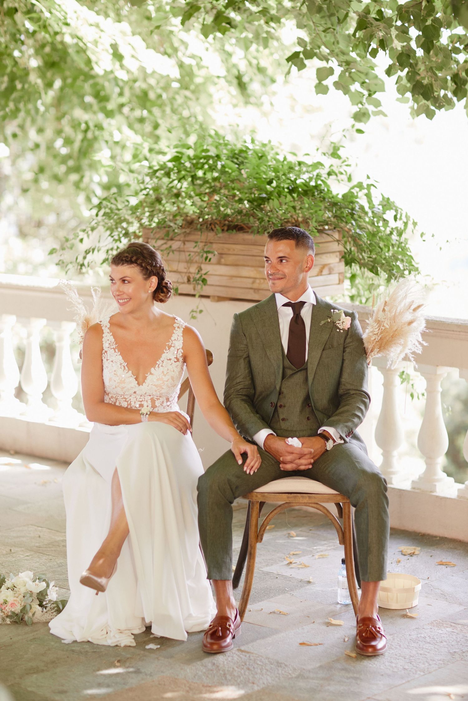 Bride and groom smiling together seated in wooden chairs during sunny outdoor wedding ceremony.