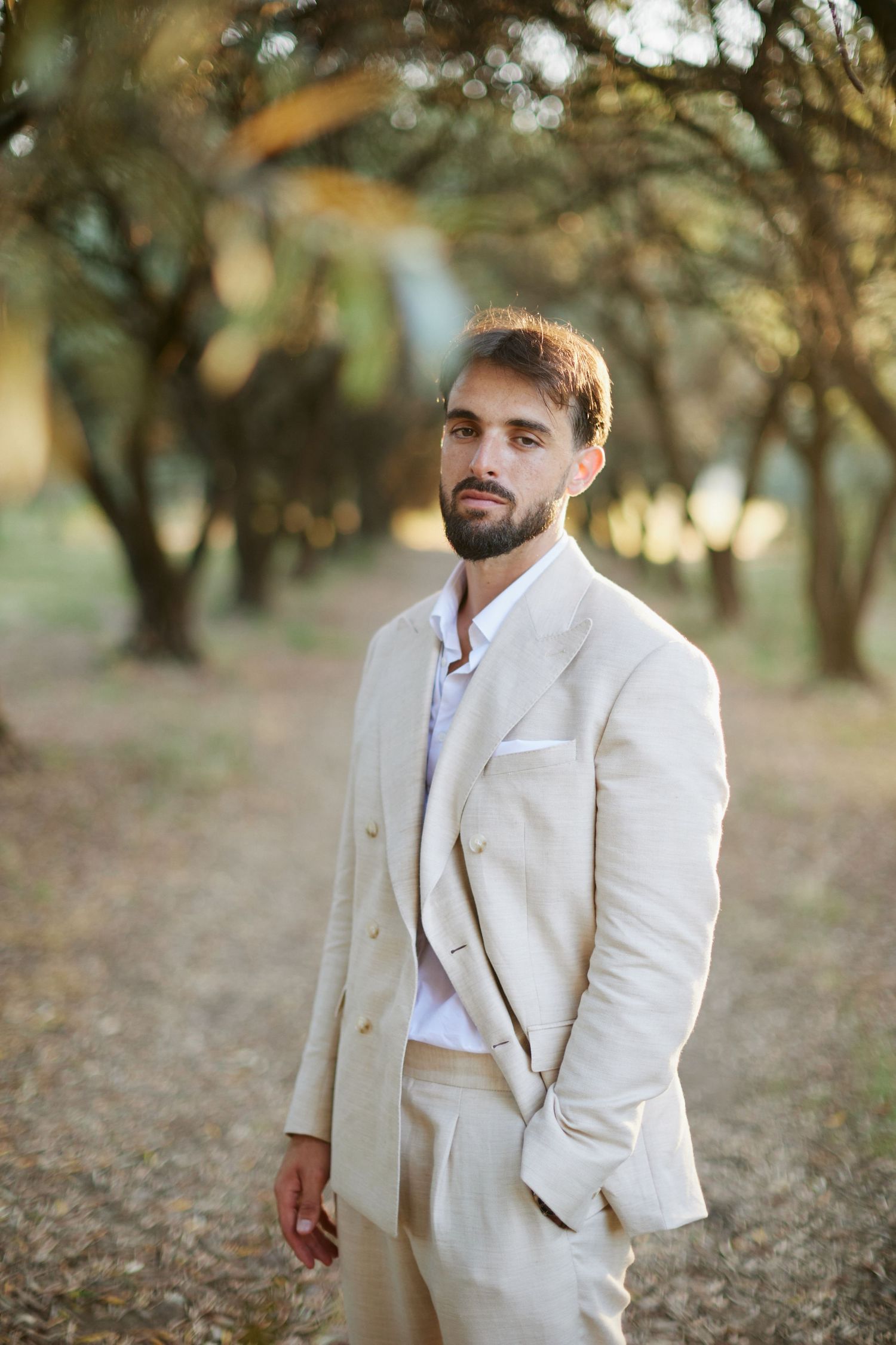 Stylish groom in cream three-piece linen suit posing among golden autumn trees in an outdoor setting.