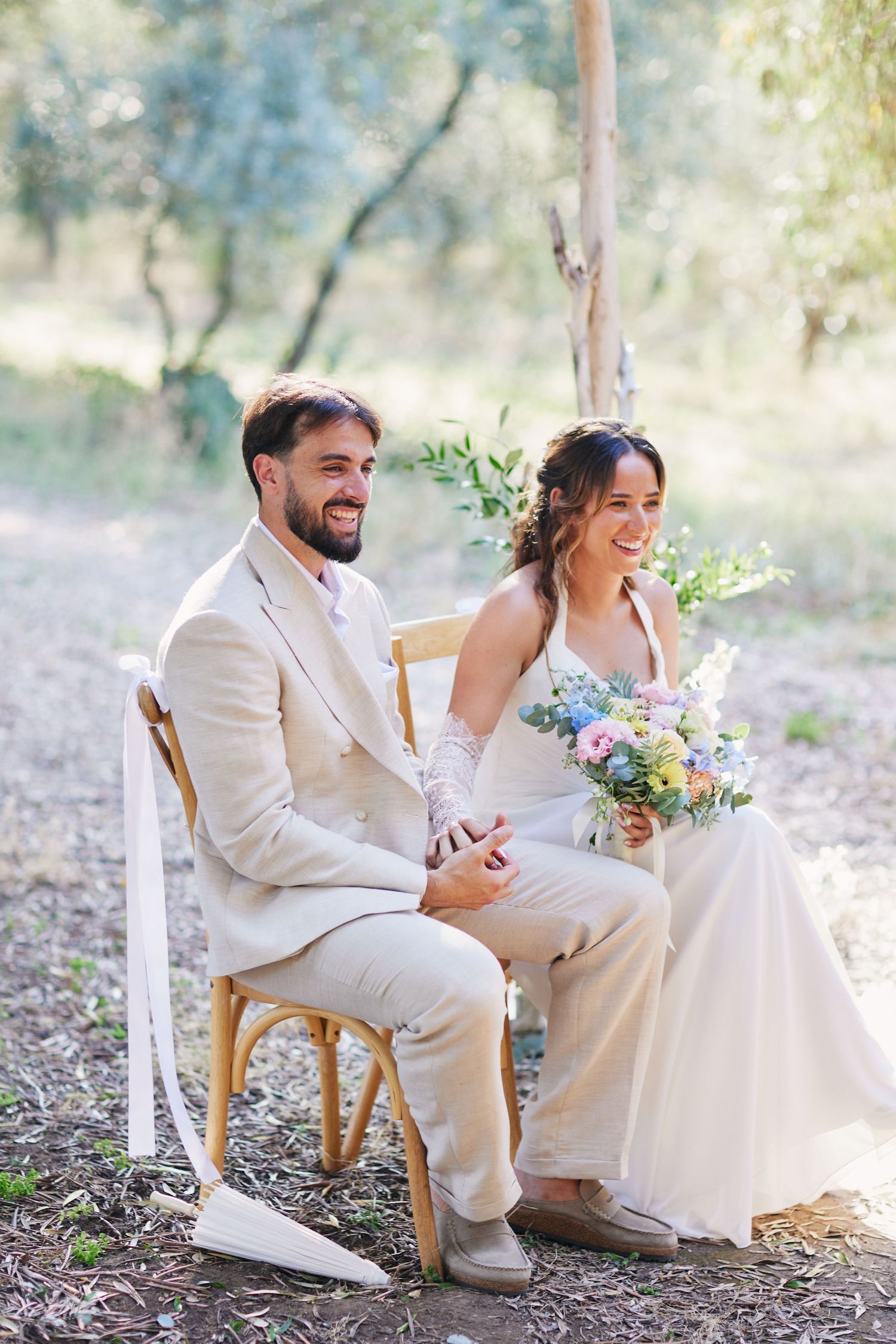 Smiling bride and groom sitting on wooden chairs outdoors during a woodland wedding ceremony.