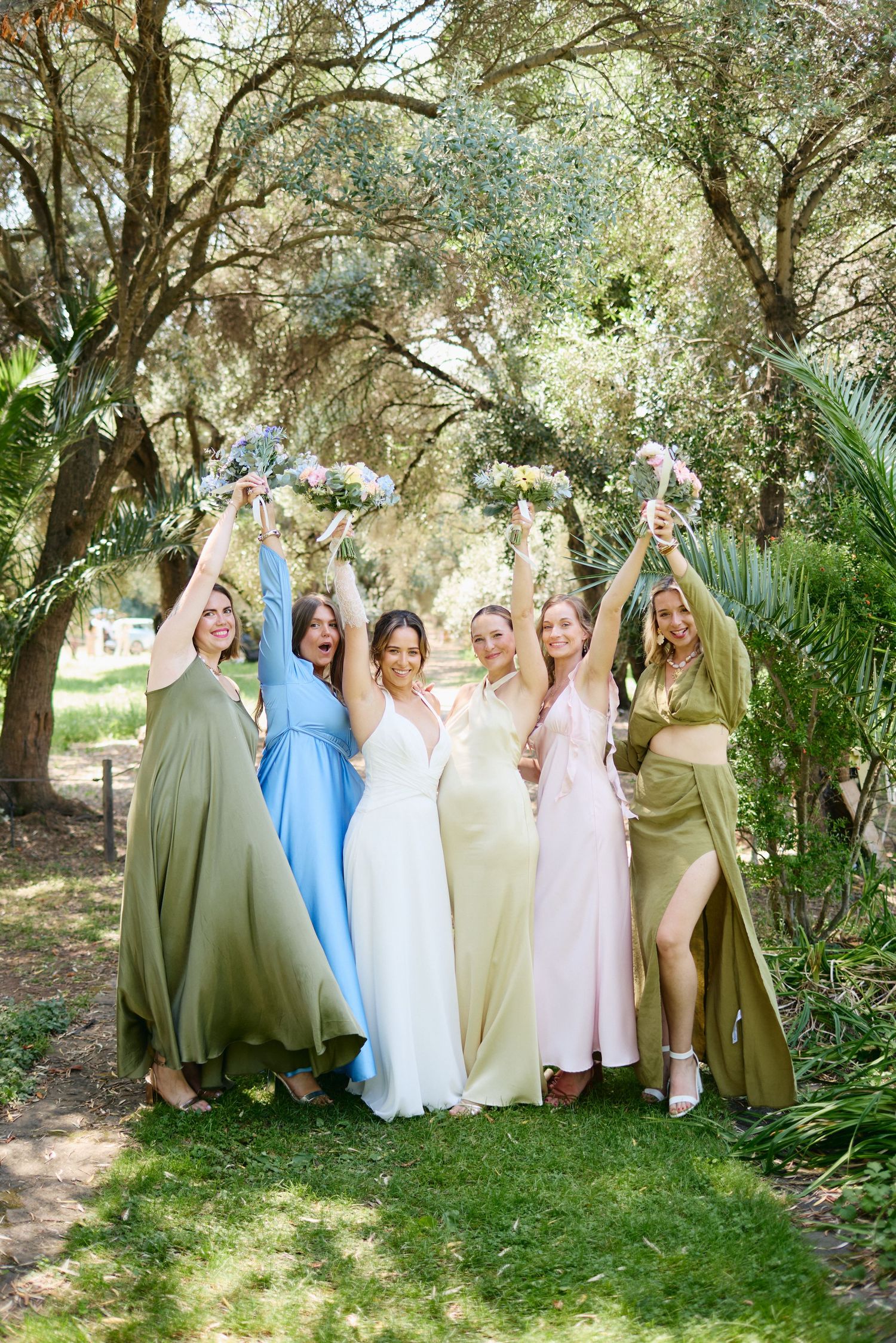Bride and five bridesmaids in mismatched pastel gowns celebrate outdoors raising bouquets under lush green trees.