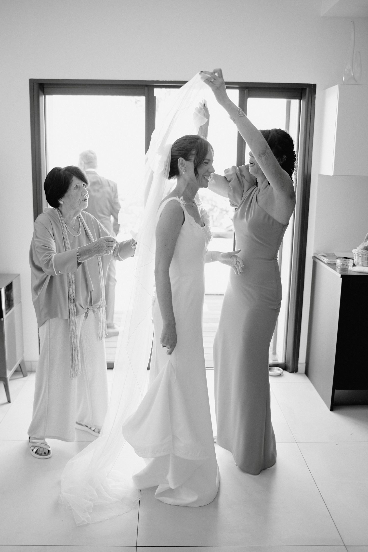 Black and white photo of bride having veil placed by bridesmaid while mother assists nearby.