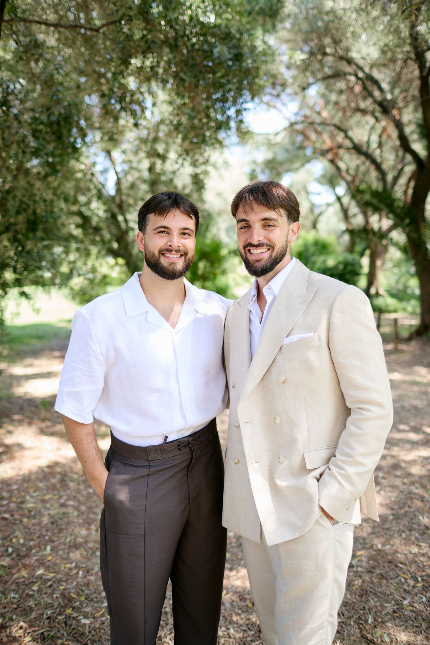 Two smiling men in white shirt and beige suit posing together outdoors in a lush green park.