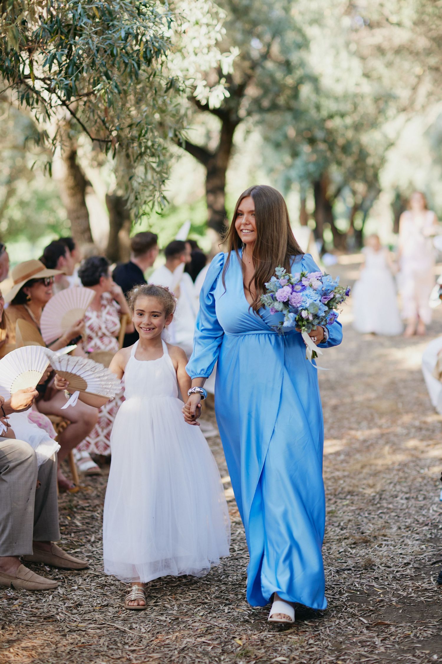 Bridesmaid in a blue gown holding a purple bouquet walks down the aisle with a flower girl in white.