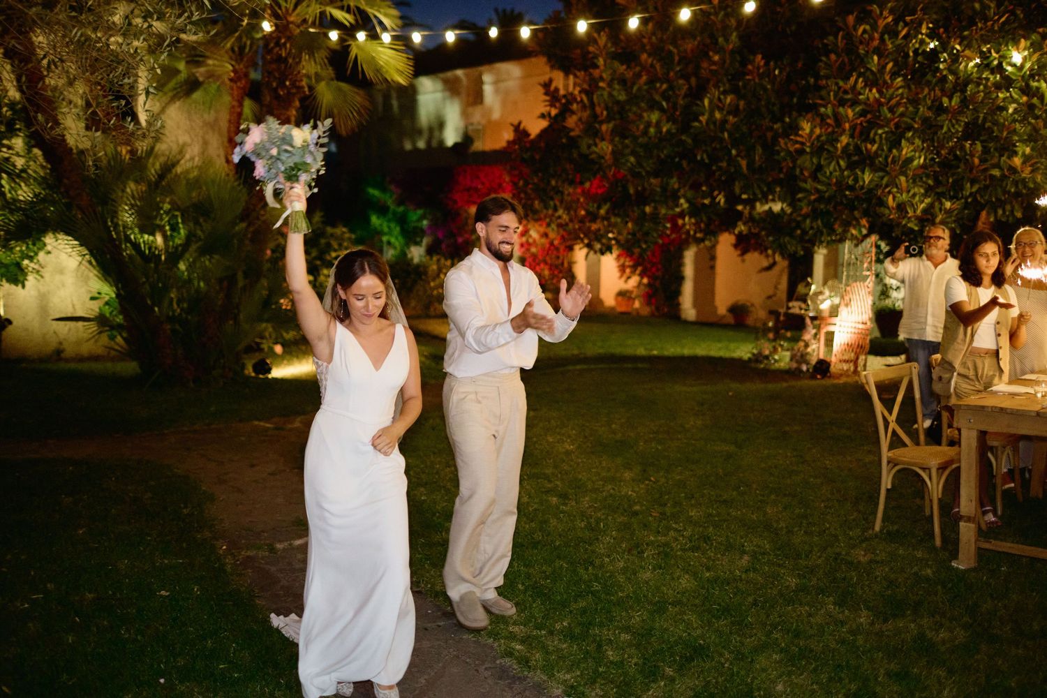 Bride and groom celebrating at night under string lights in lush garden wedding venue.