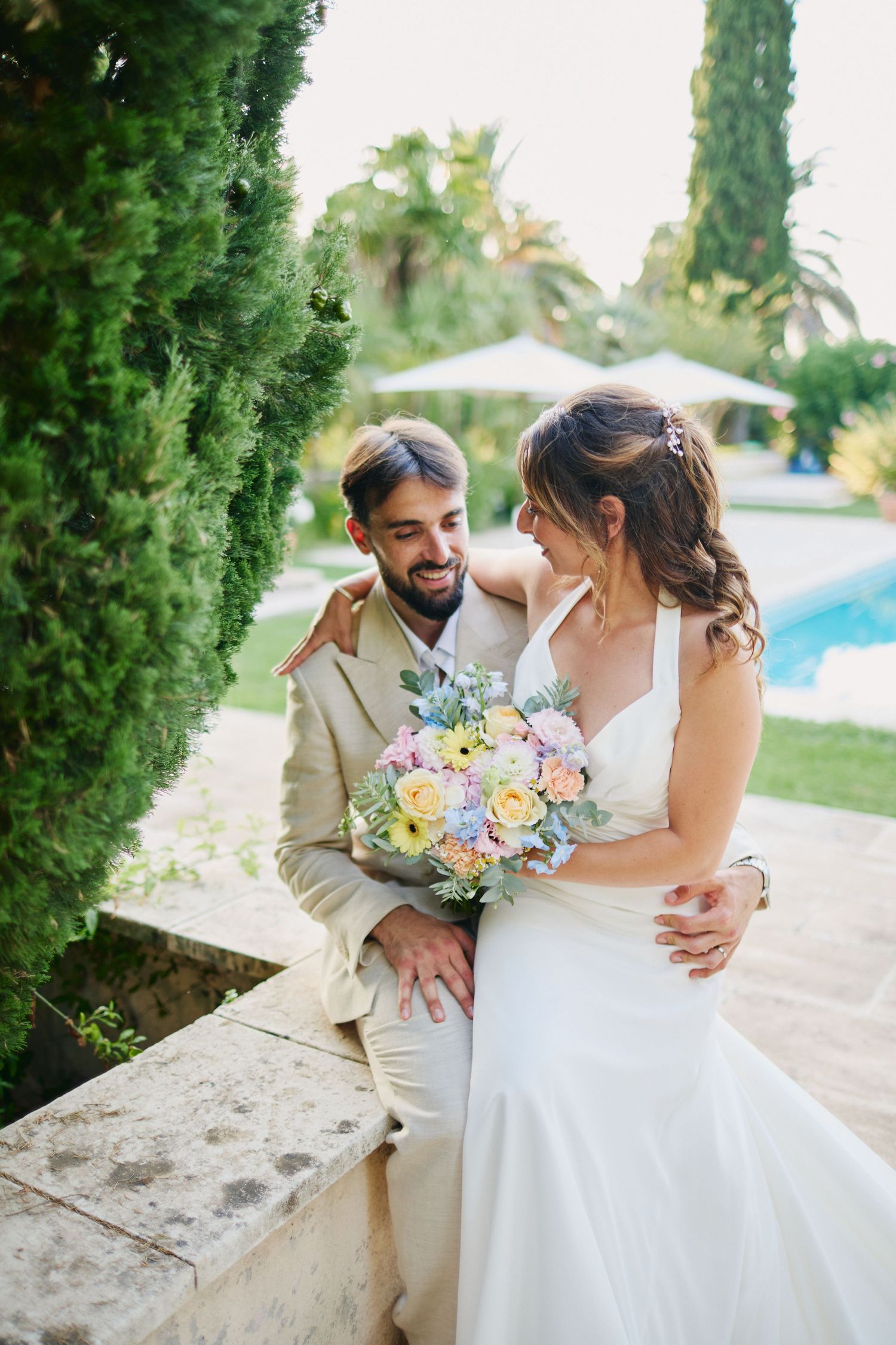 Bride and groom embracing near pool in garden, holding pastel wedding bouquet.