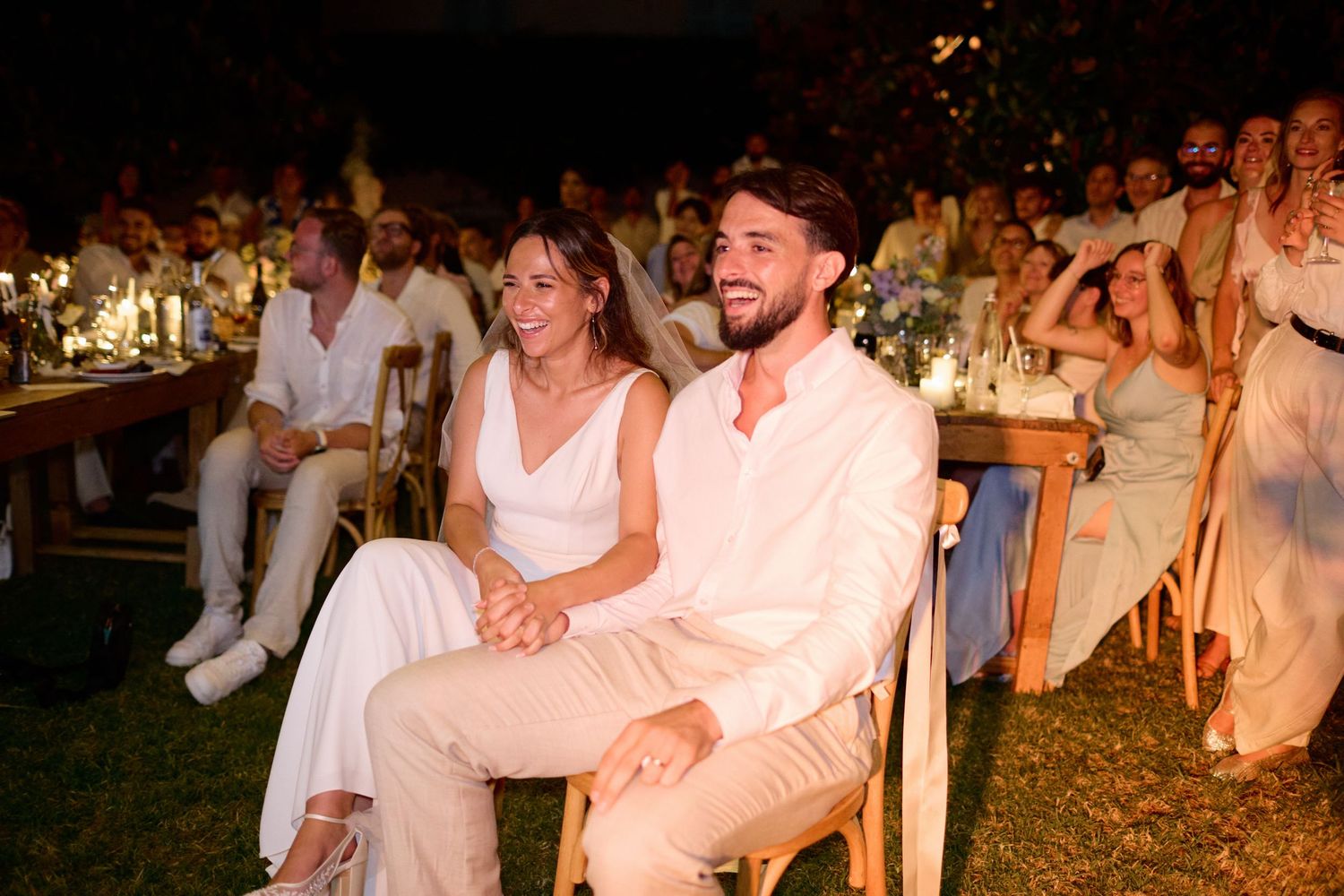 Happy bride and groom seated together at outdoor wedding reception surrounded by guests in white attire.