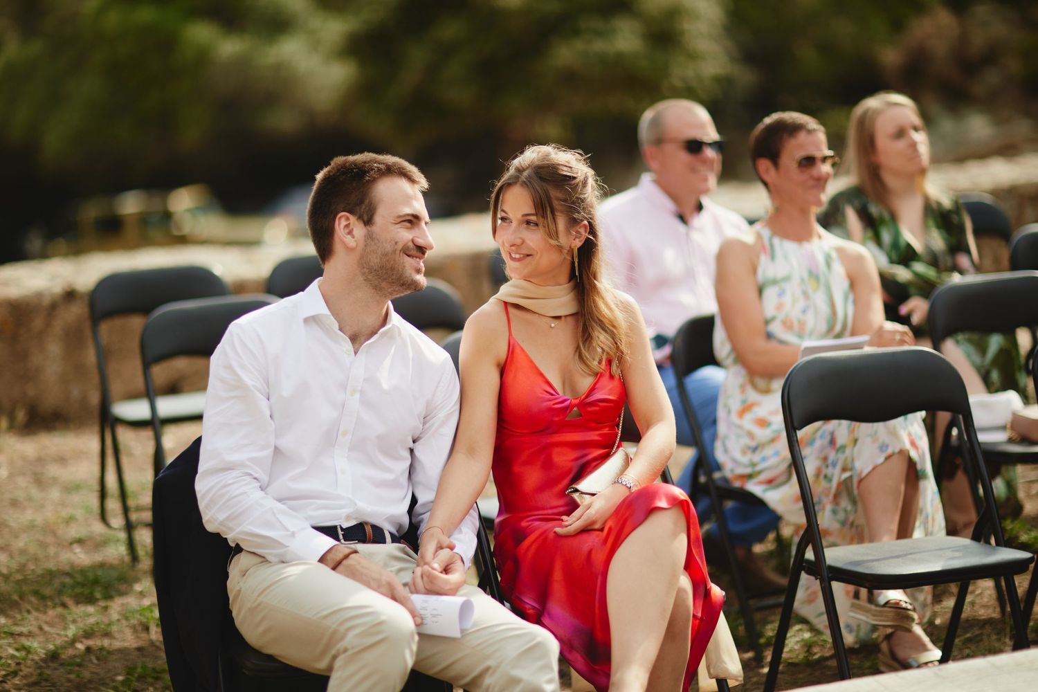 Smiling couple, woman in red dress and man in white shirt, seated as wedding guests at outdoor ceremony.