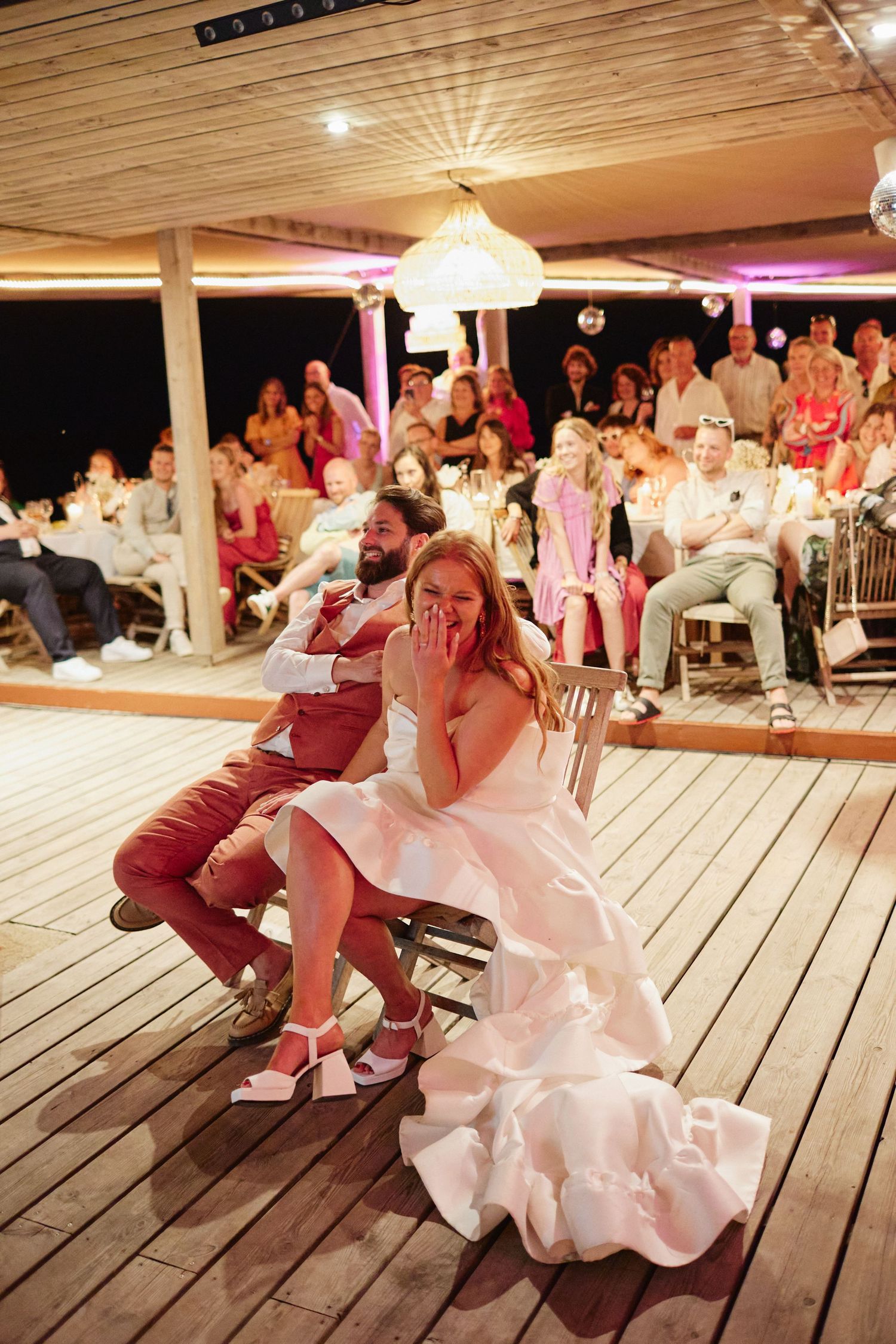 Bride and groom sitting on dance floor laughing during fun wedding reception game with guests watching.