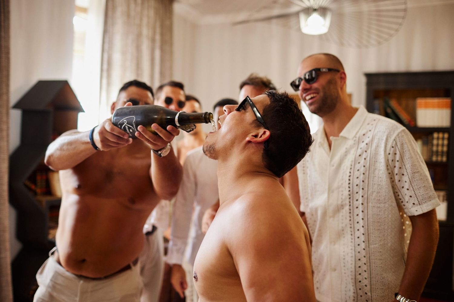 Groomsman drinking from a bottle gun while friends laugh and celebrate during pre-wedding preparations indoors.