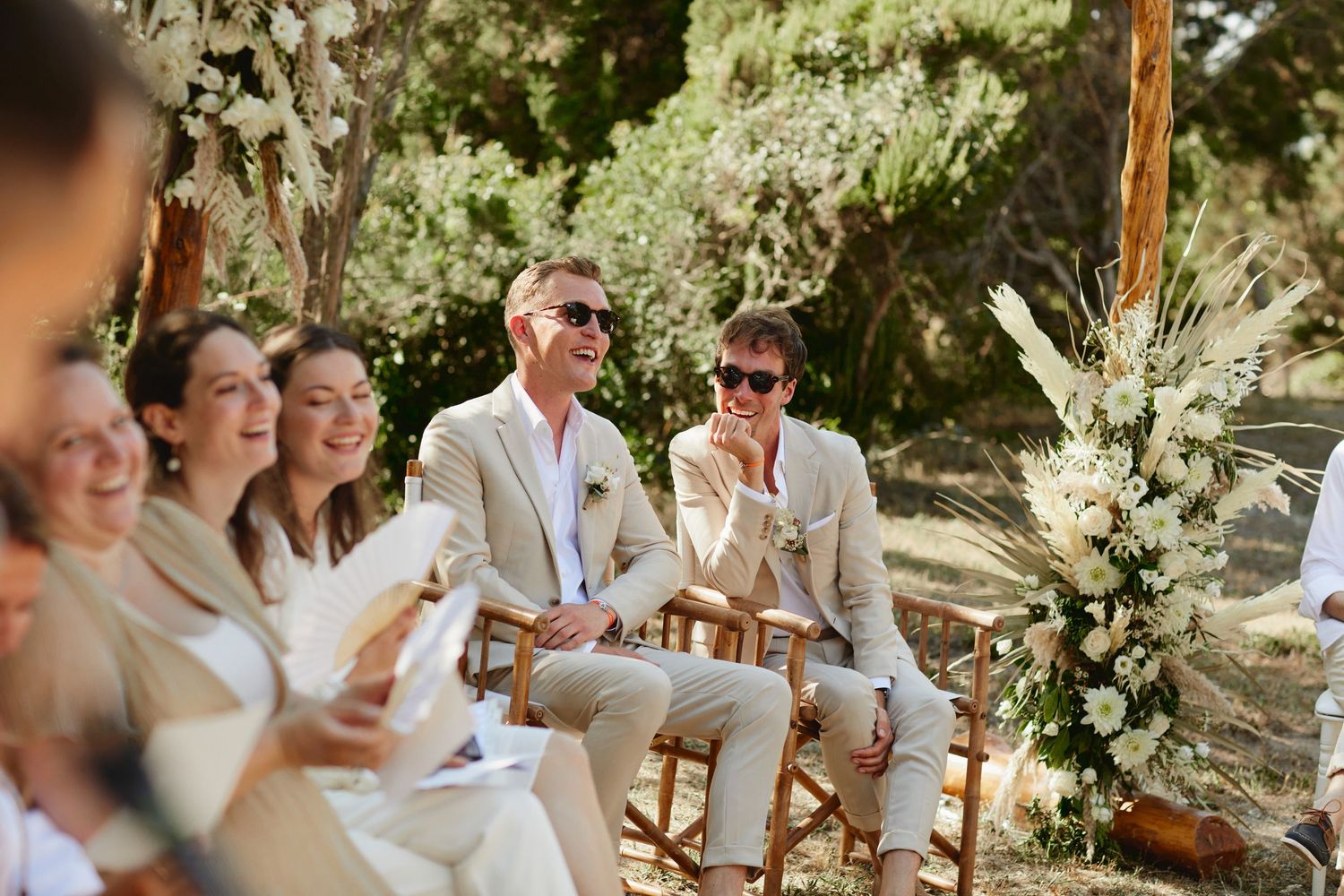 Two grooms in beige suits laugh joyfully while seated among guests at an outdoor wedding.