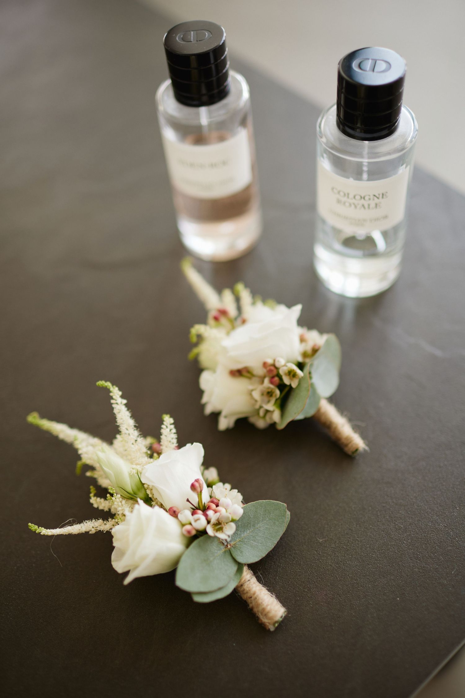 Two white floral boutonnieres with eucalyptus beside Dior Cologne Royale perfume bottles on gray surface.