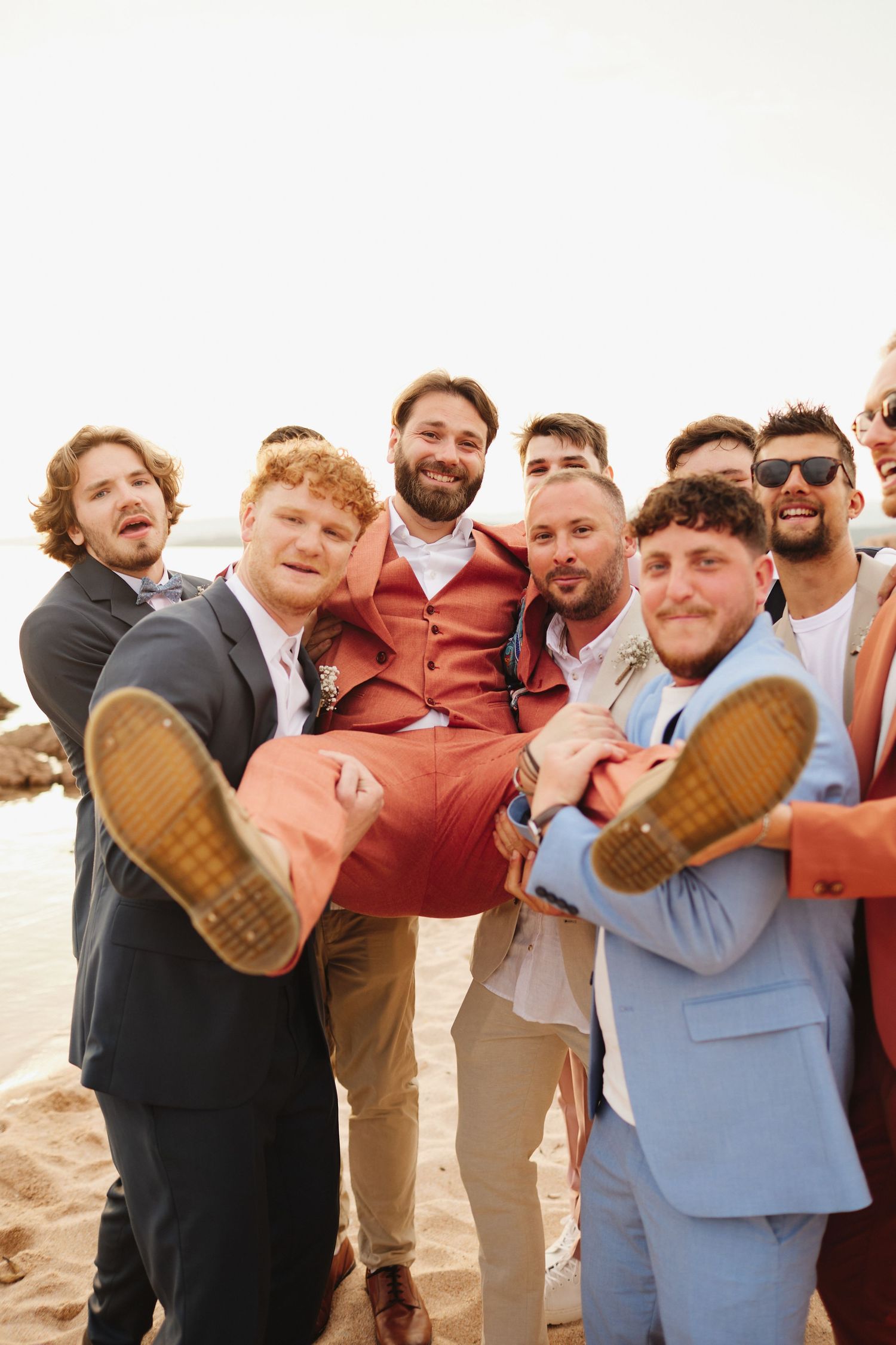 Groomsmen carrying smiling groom in coral suit on beach during fun wedding party photo.