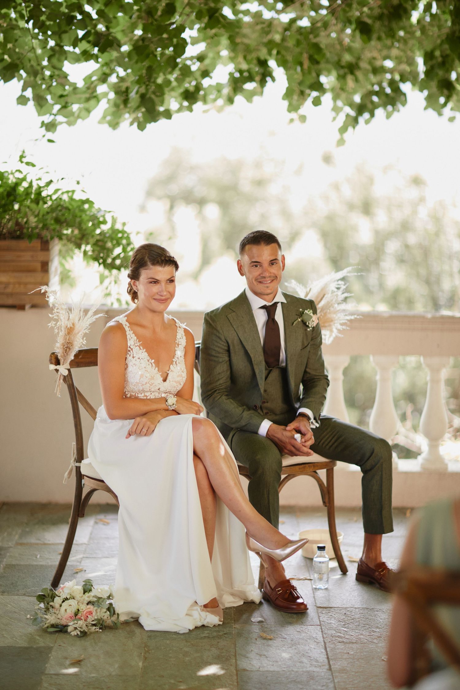 Bride in lace wedding dress and groom in olive suit smiling during outdoor ceremony.