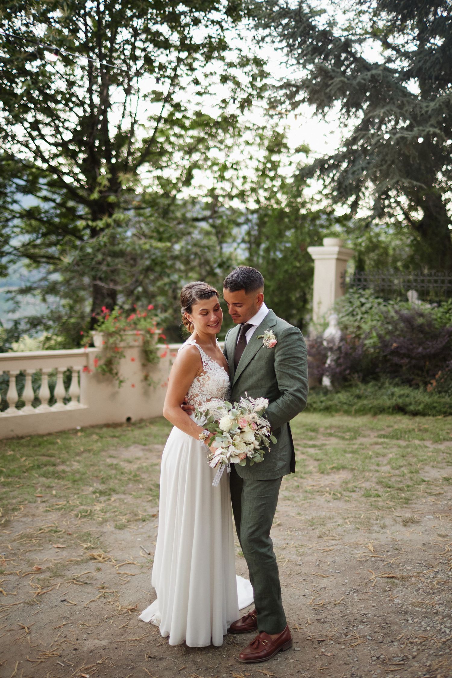 Wedding couple embrace outdoors, bride in elegant white gown and groom in green suit holding a floral bouquet.