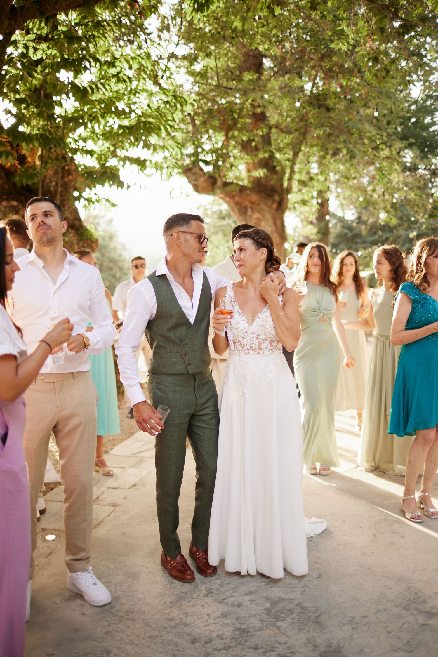 Bride and groom walk hand in hand among smiling guests at an outdoor wedding celebration.
