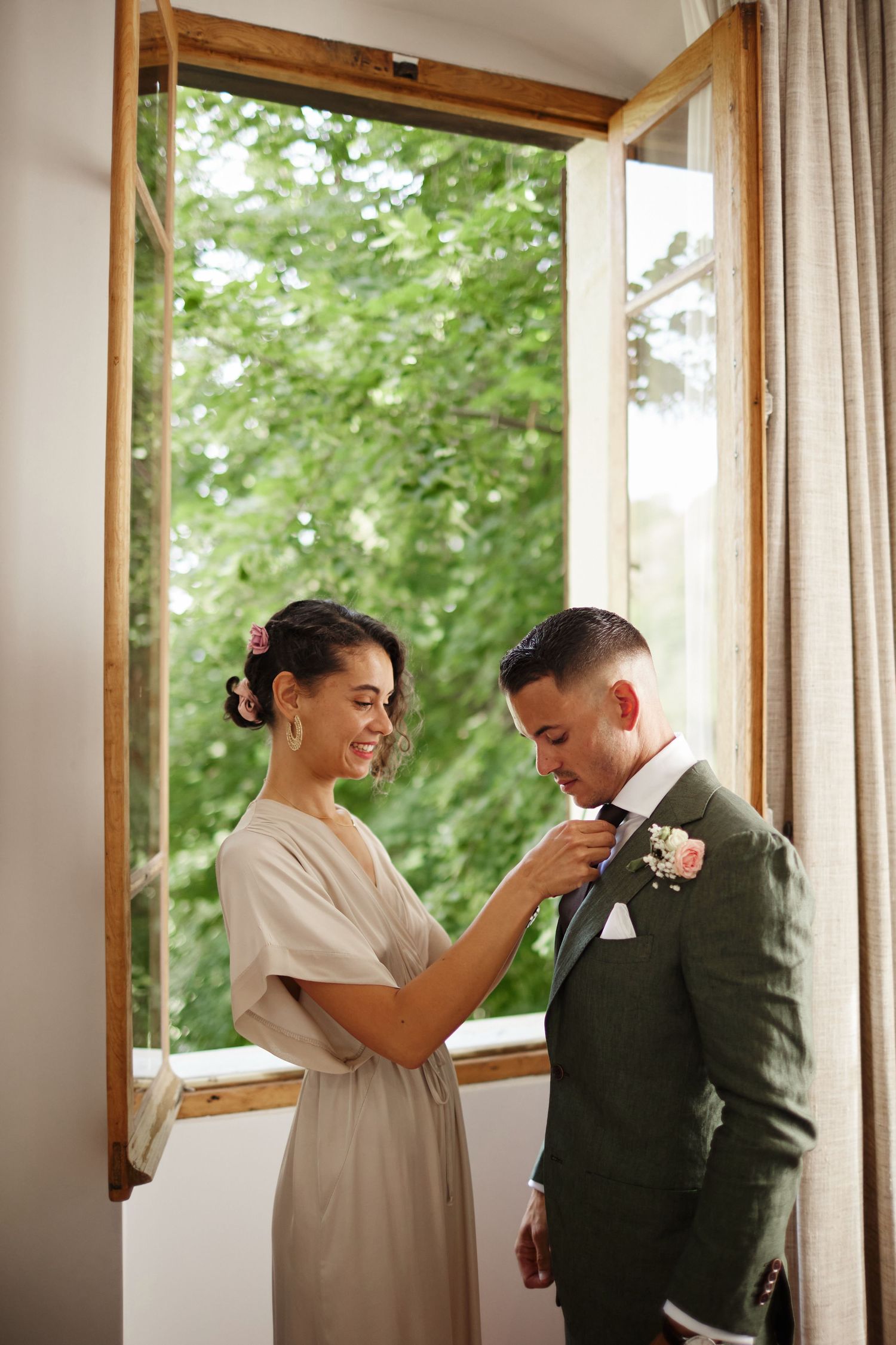 Smiling bridesmaid in a beige dress pinning a boutonniere on the groom in a green suit by an open window.