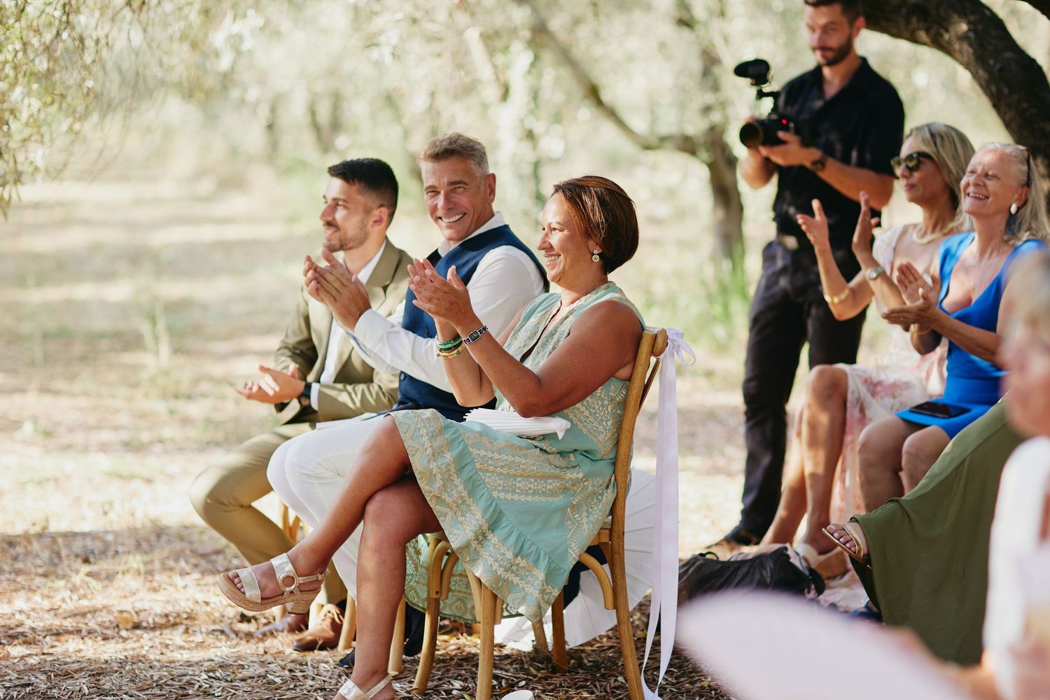 Wedding guests clap and celebrate during outdoor ceremony seated on wooden chairs under olive trees.