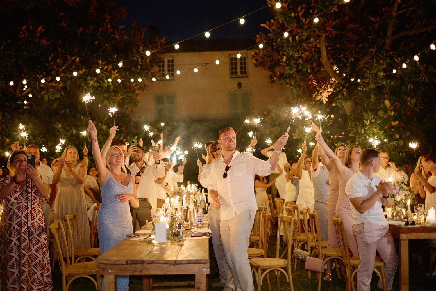 Wedding guests celebrating with sparklers under string lights at elegant outdoor nighttime reception.
