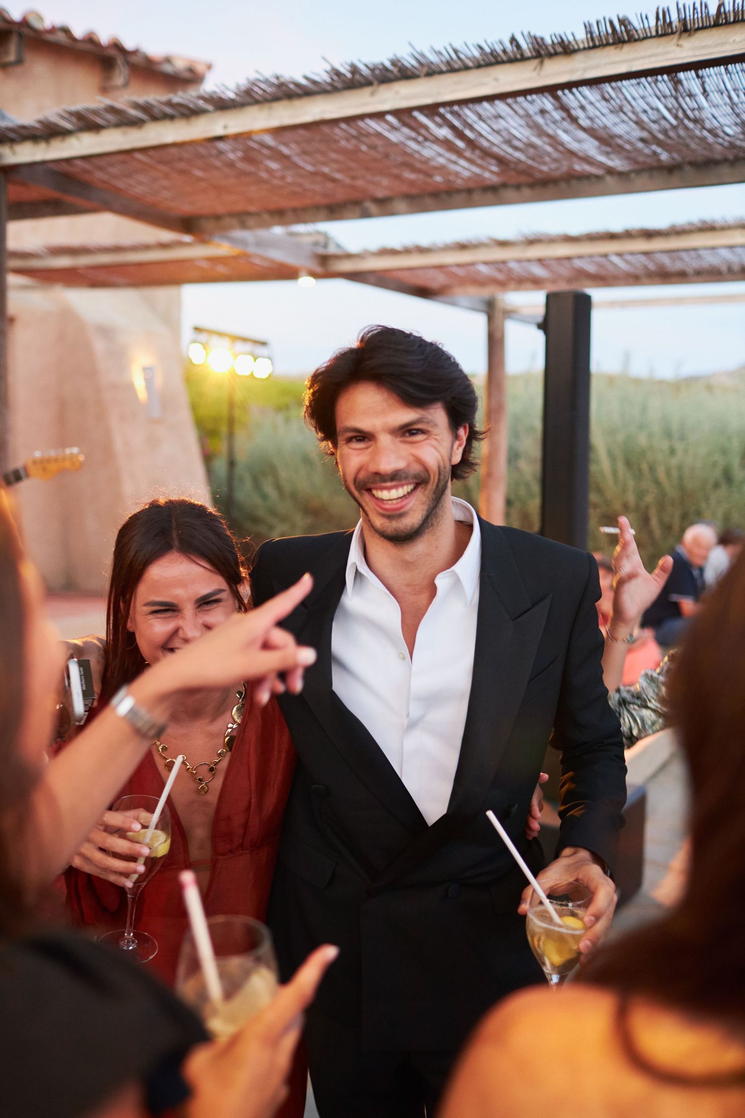 Smiling man in black suit holds cocktail at elegant outdoor wedding reception party.