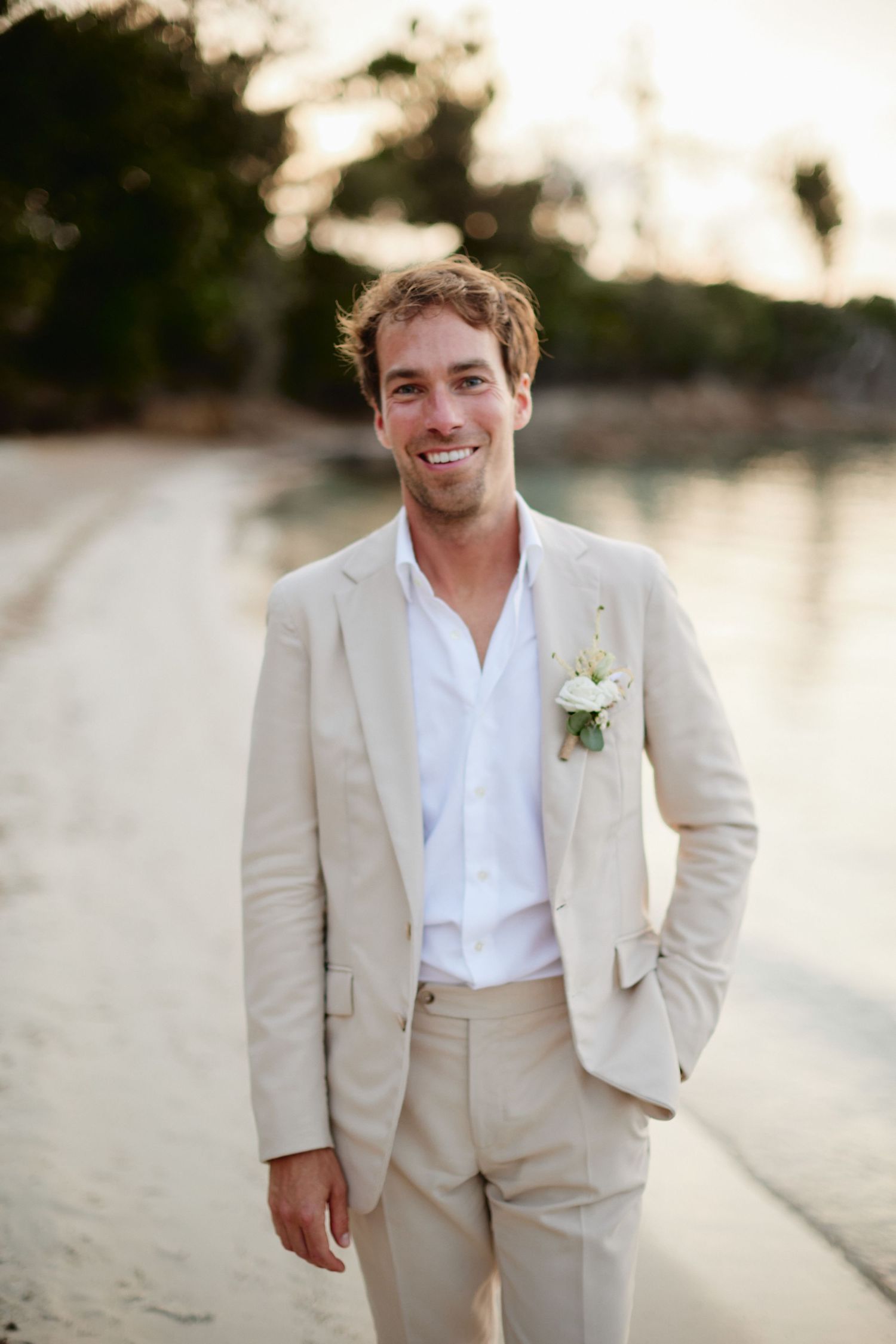 Smiling groom in beige linen suit with white boutonniere standing on a tropical beach.