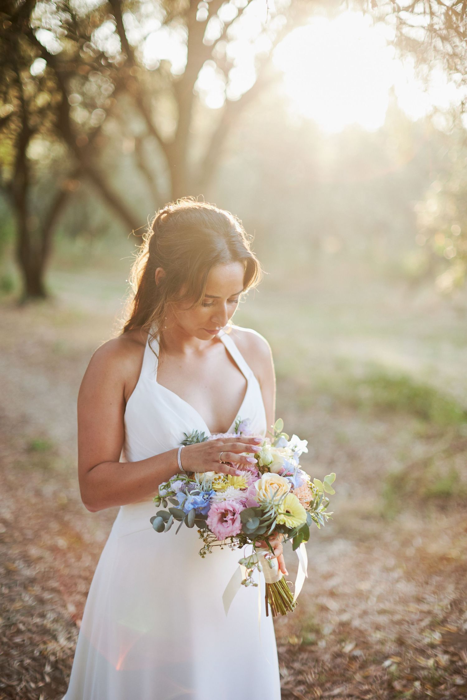 Bride in white halter dress gazing at a pastel floral bouquet in a sunlit outdoor grove setting.