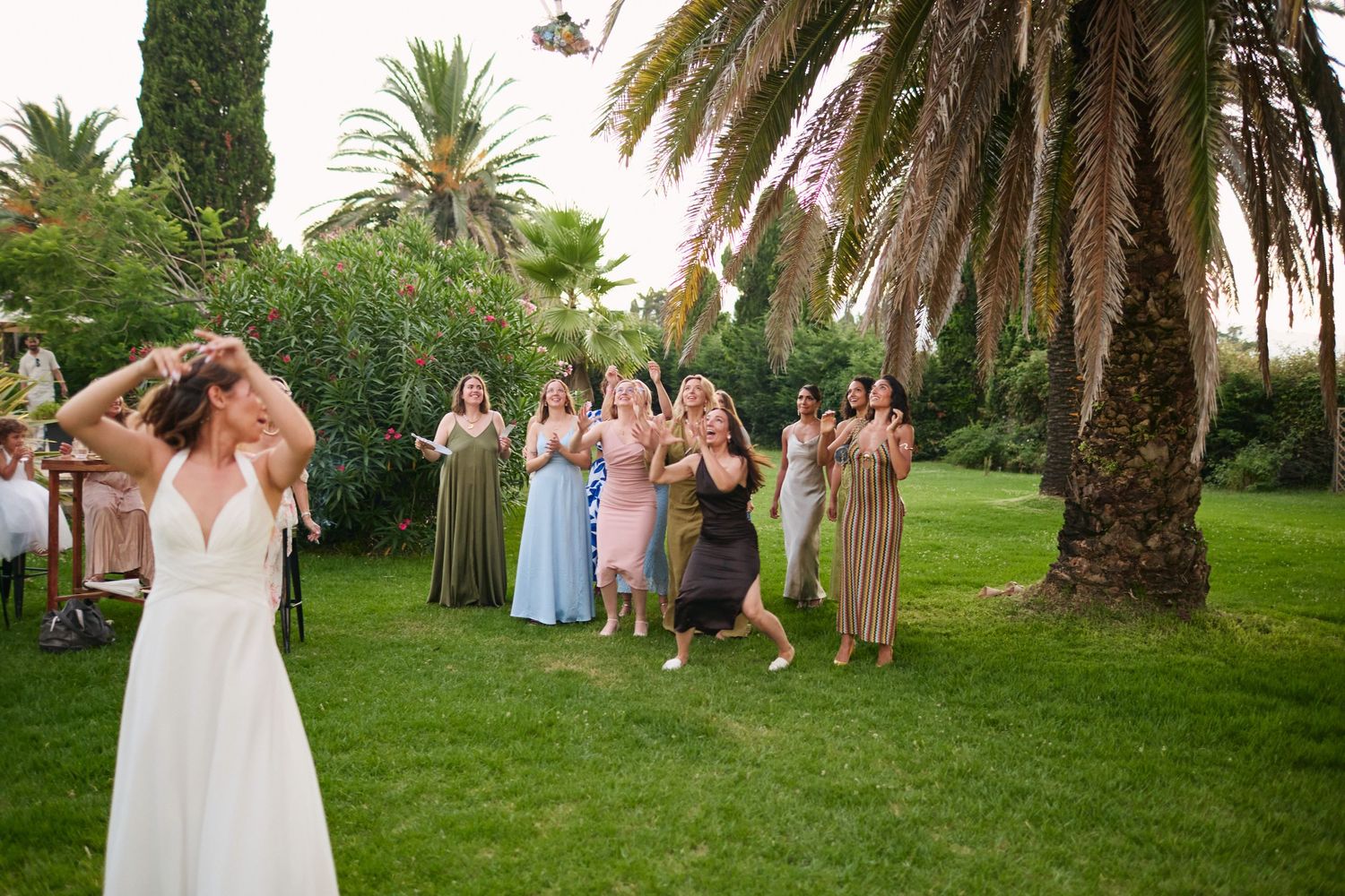 Bride in white gown tossing bouquet to excited guests in colorful dresses on lush green lawn.