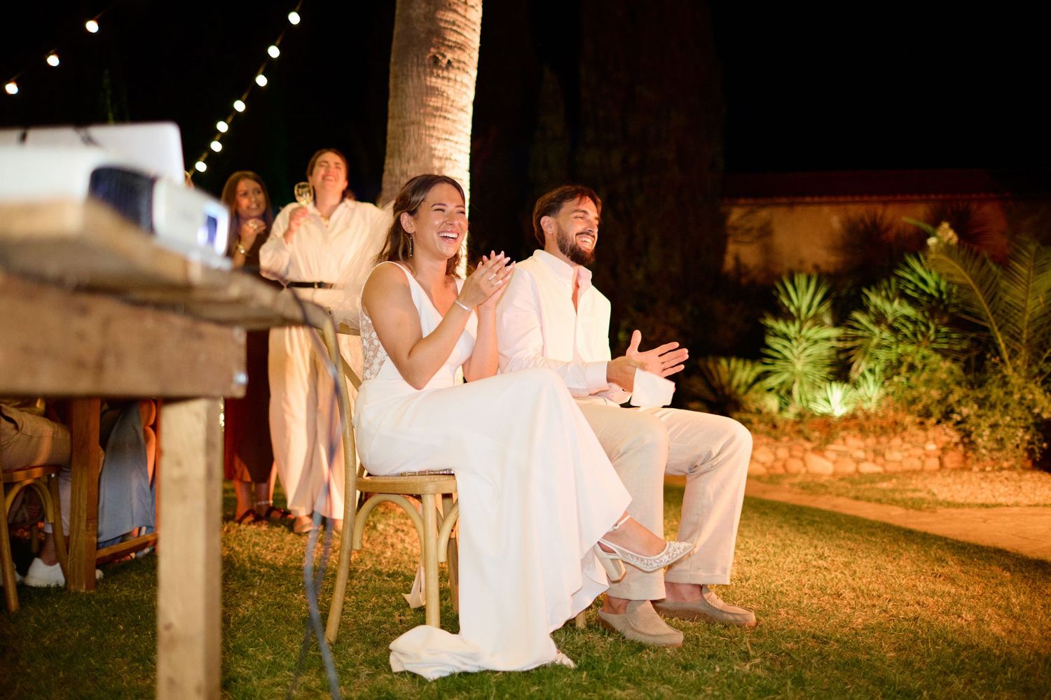 Smiling bride and groom sit together outdoors at their evening wedding reception under string lights.