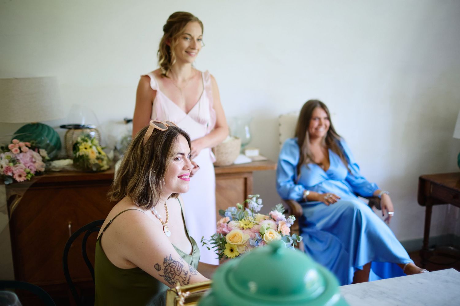 Bridesmaids laughing and relaxing indoors during wedding morning preparations with colorful bouquets.